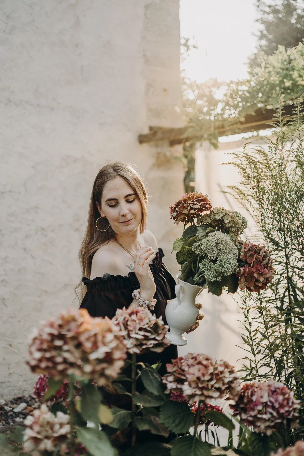 Jeune femme avec un haut noir à manches courtes, portant des boucles d'oreilles en or, tenant une cruche blanche avec des fleurs dans un jardin, en train de regarder les fleurs.