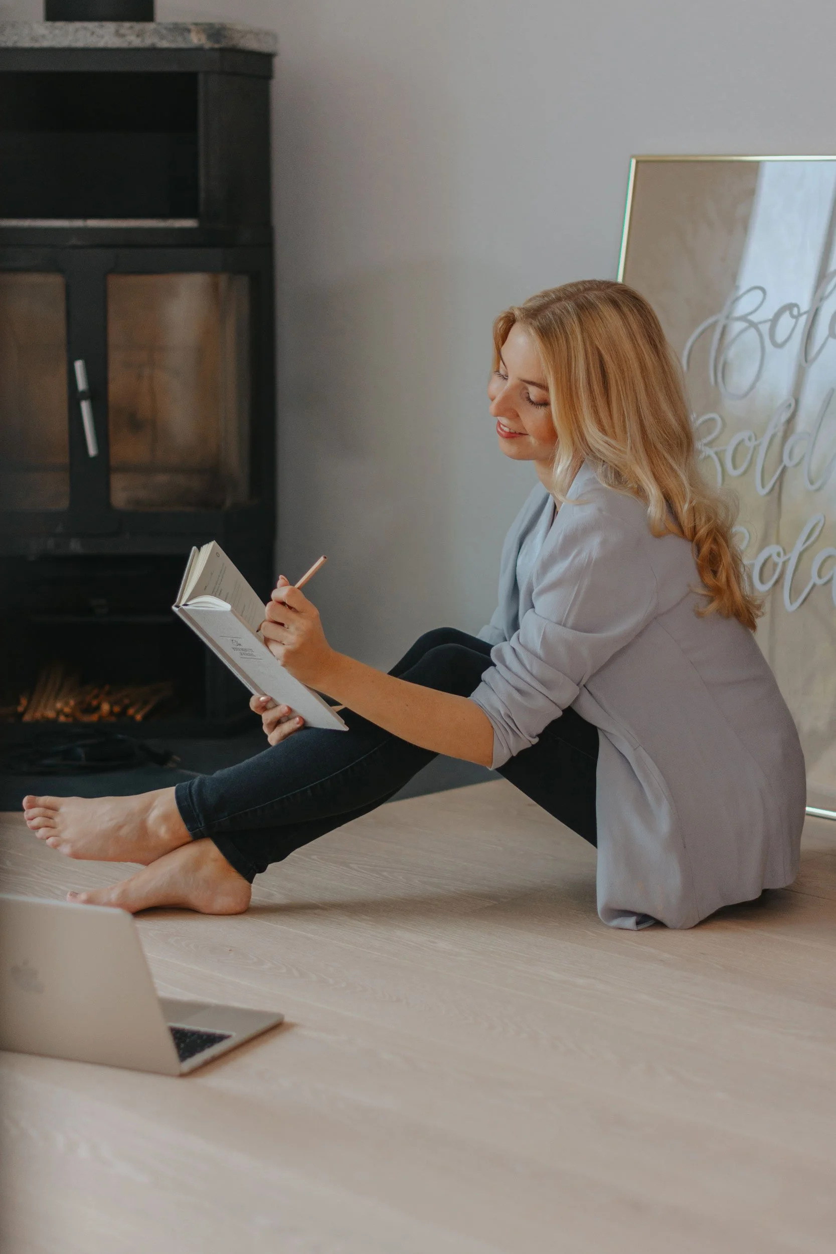 Femme assise par terre, lisant un livre et souriant, avec un ordinateur portable devant elle, dans un intérieur moderne.