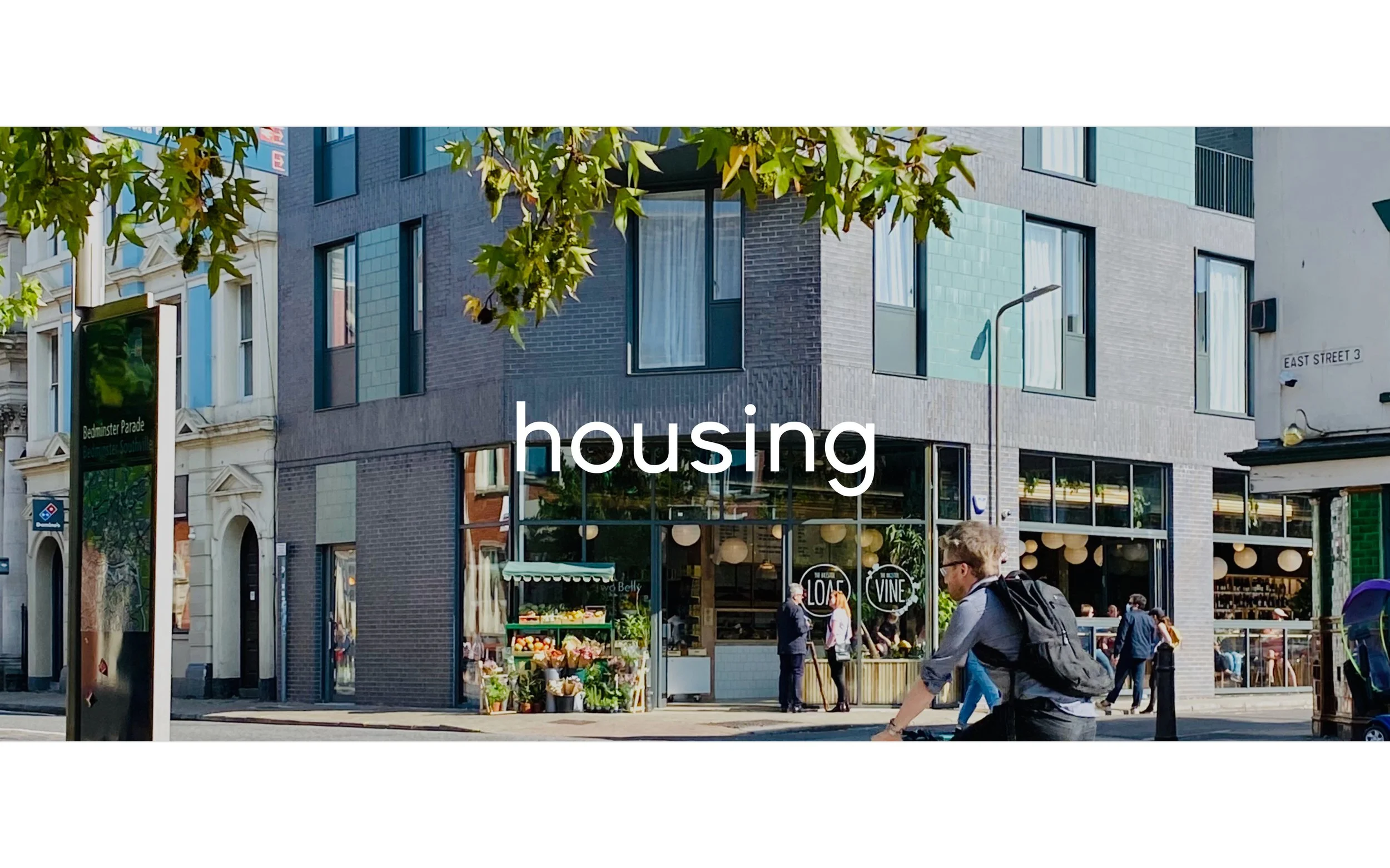 Barefoot Architects Ltd — Urban street scene with modern apartment building, small grocery stand with produce, pedestrians including a cyclist carrying a backpack, and street s