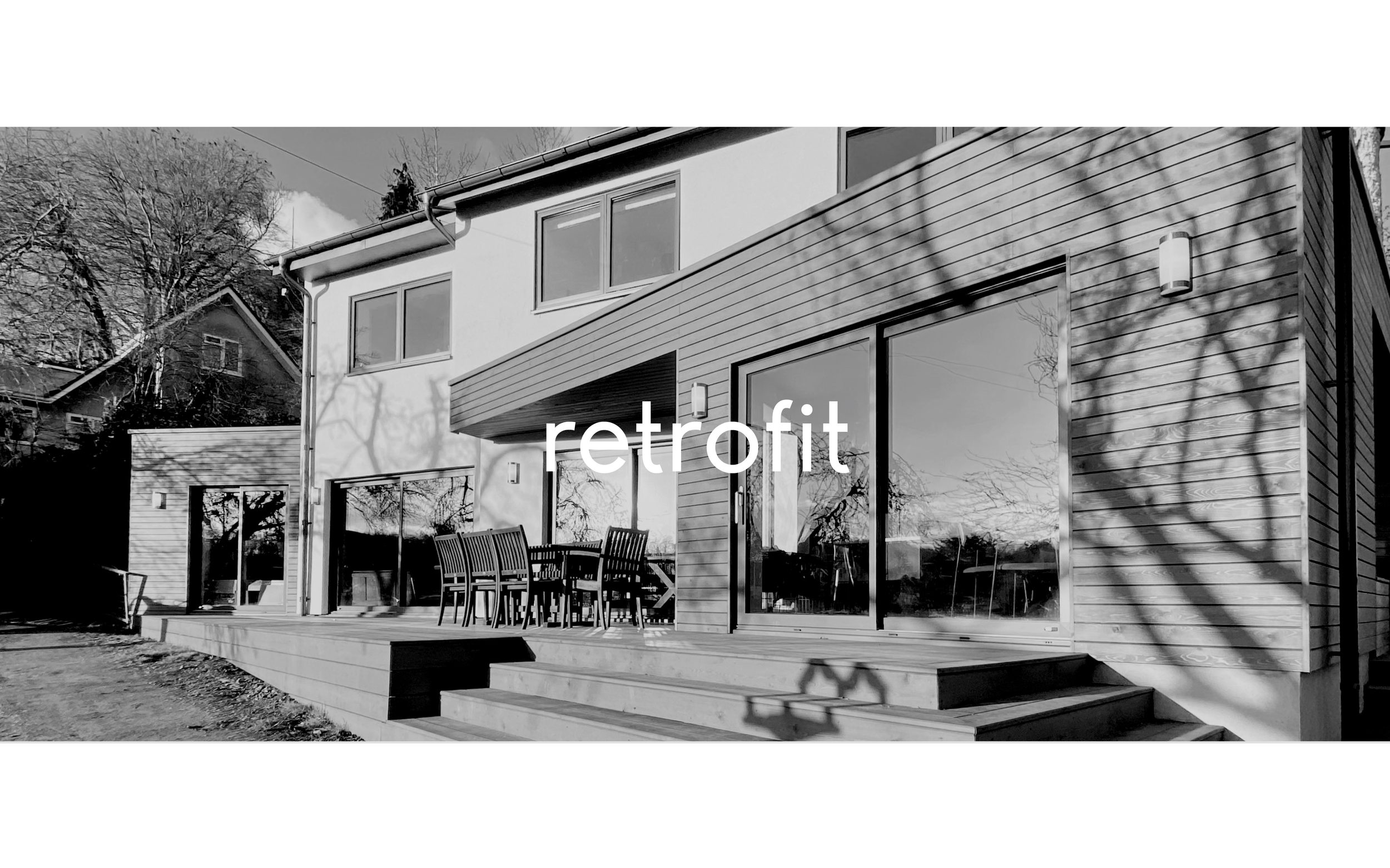 Barefoot Architects Ltd — Modern house with a wooden deck, outdoor dining table, large sliding glass doors, and shadows of trees on the exterior wall.