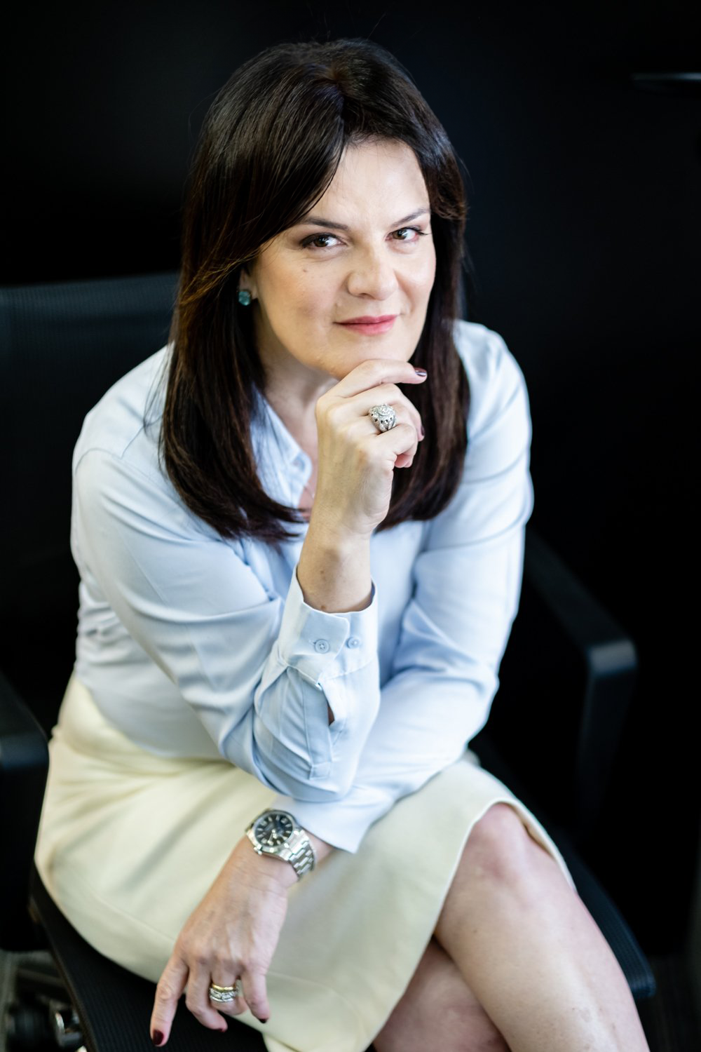 Mulher com cabelo castanho escuro, vestida com camisa azul e saia bege, sentada em cadeira preta, com uma expressão confiante, olhando para a câmera, vestindo acessórios de joias e relógio.