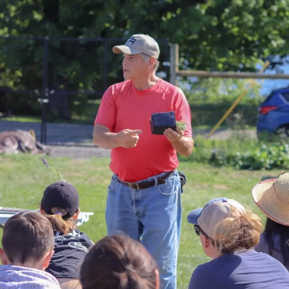 A man in a red shirt and cap holding a small black basket with plants, speaking to a group of children and adults outdoors on a sunny day.