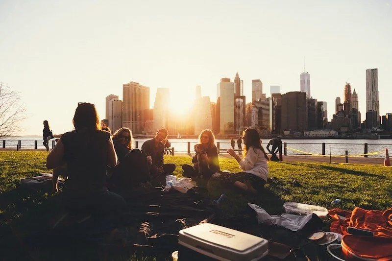 A group of friends sits on a grassy hill having a picnic, silhouetted against a bright sunset and the Manhattan skyline.