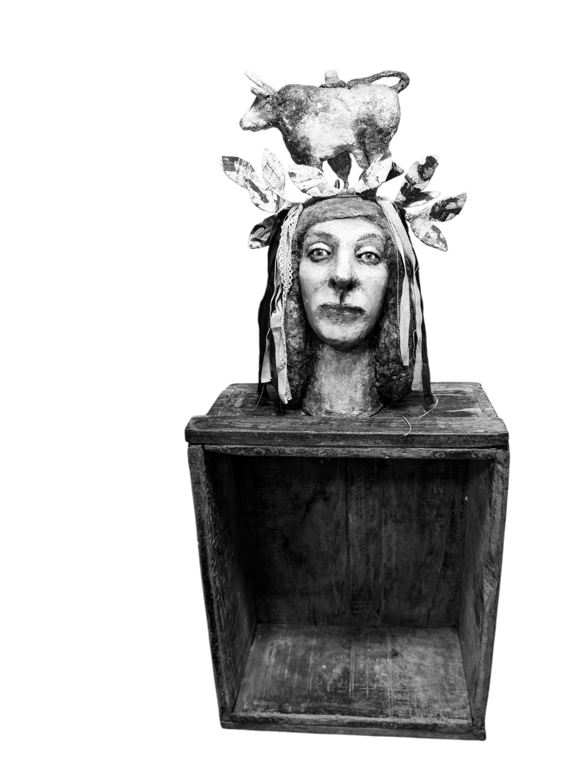 A sculpture featuring a human face with an elaborate headpiece made of leaves and a figure of a cow on top, set on top of a wooden box.