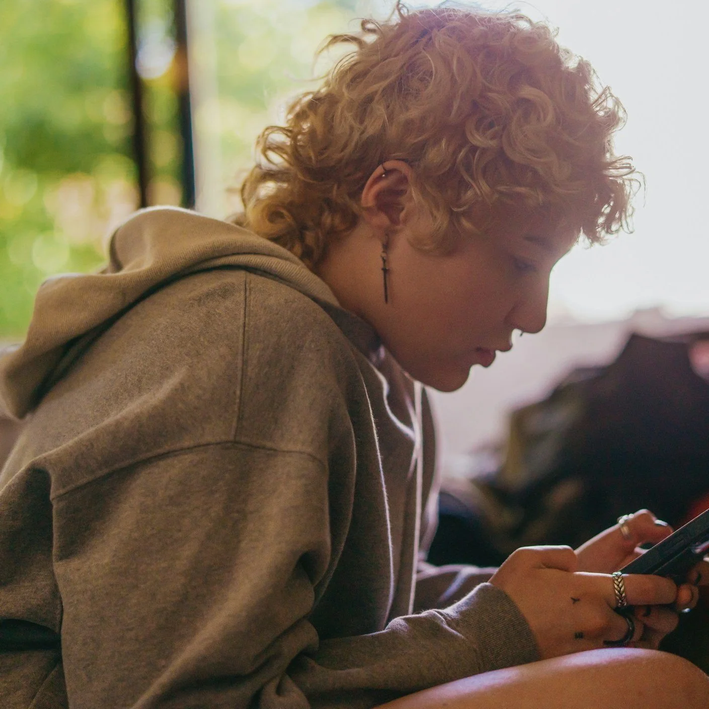 Young person with curly blond hair wearing a beige hoodie, sitting indoors and looking at a smartphone, representing individual counselling and personal growth at Black Onyx Counselling in London, Ontario
