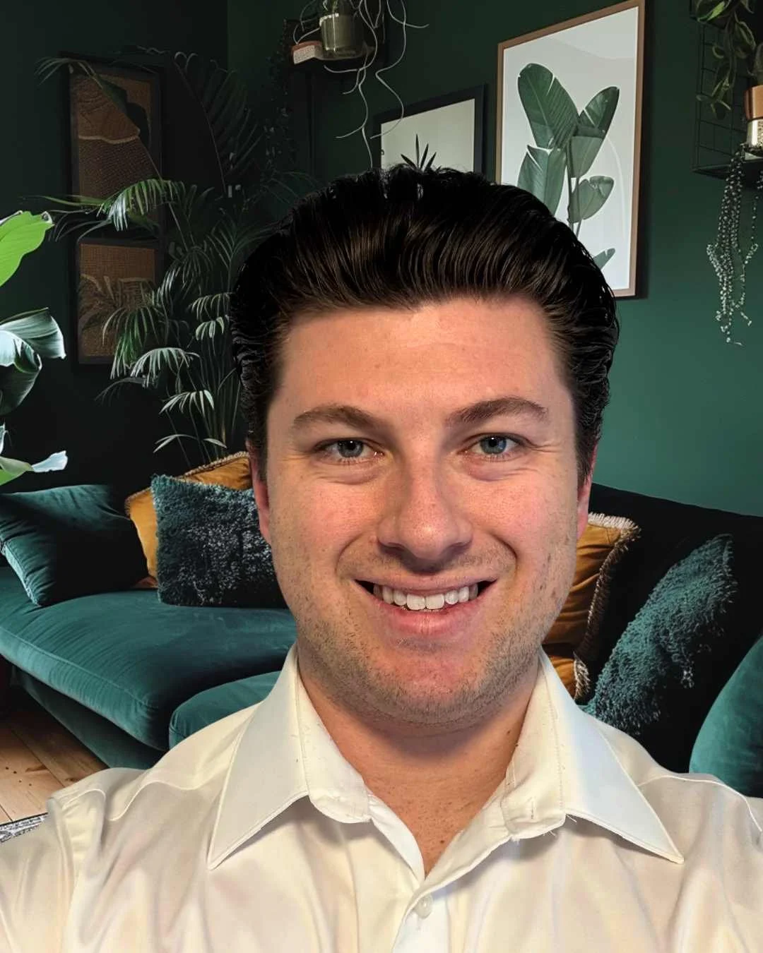 A man with dark, slicked-back hair smiling in a room decorated with green walls, framed botanical art, and lush indoor plants.