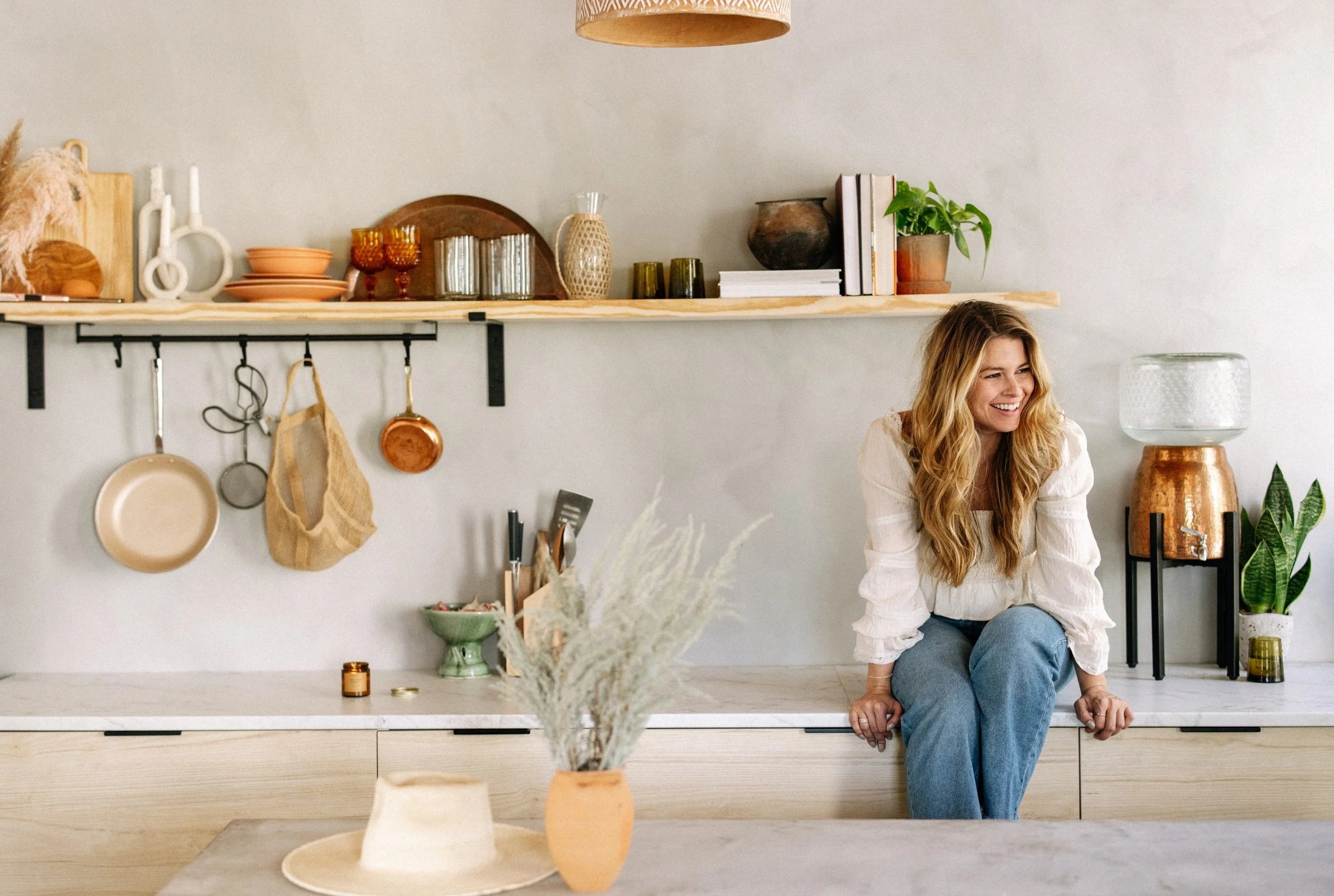 A woman with long blonde hair, wearing a white blouse and blue jeans, sitting on a kitchen counter and smiling.