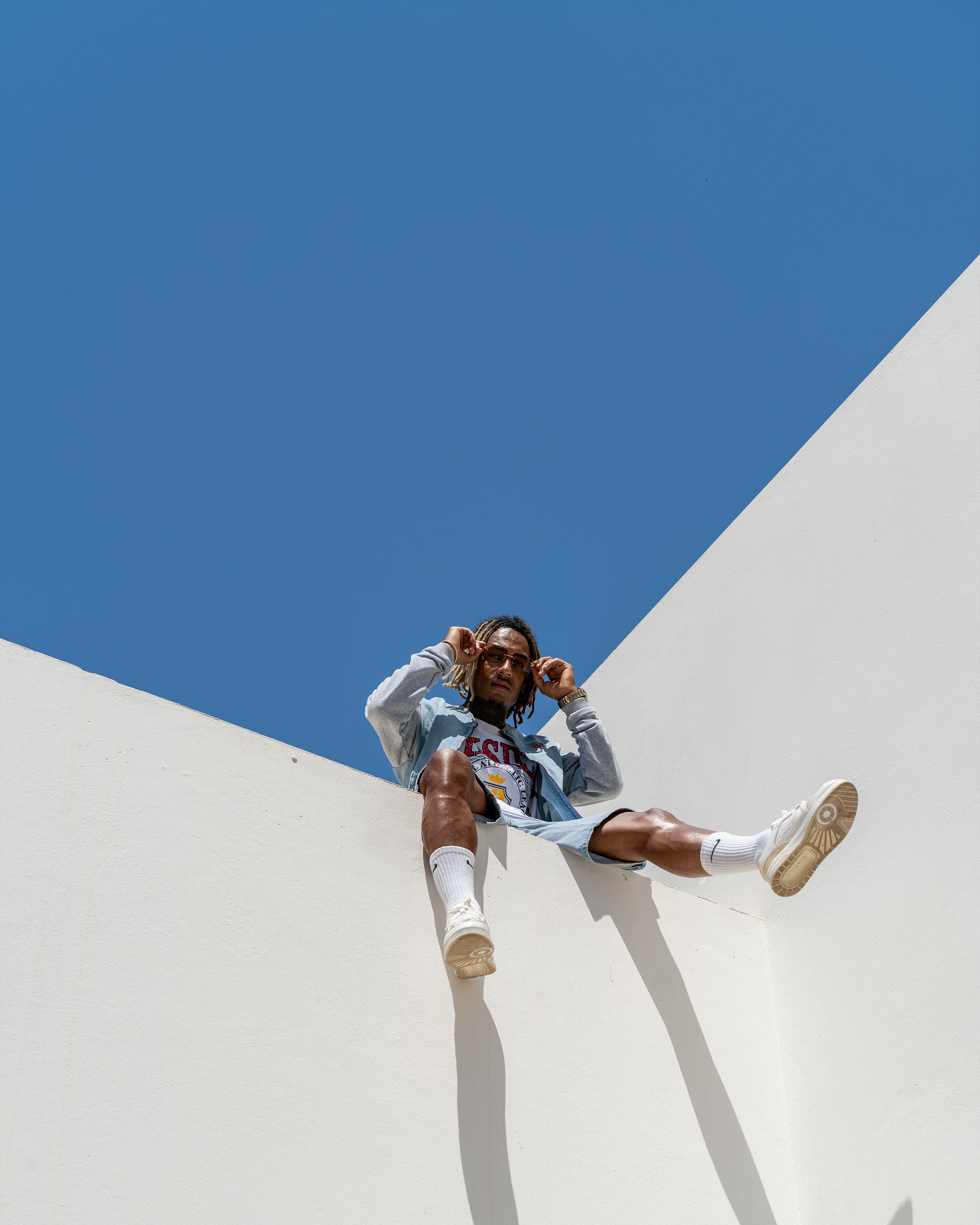 A young man with dreadlocks sitting on a white architectural structure against a clear blue sky, adjusting his sunglasses.