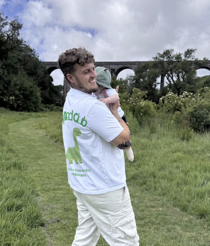 A smiling man holding a baby in a green hat outdoors on a grassy trail, with a bridge and trees in the background.