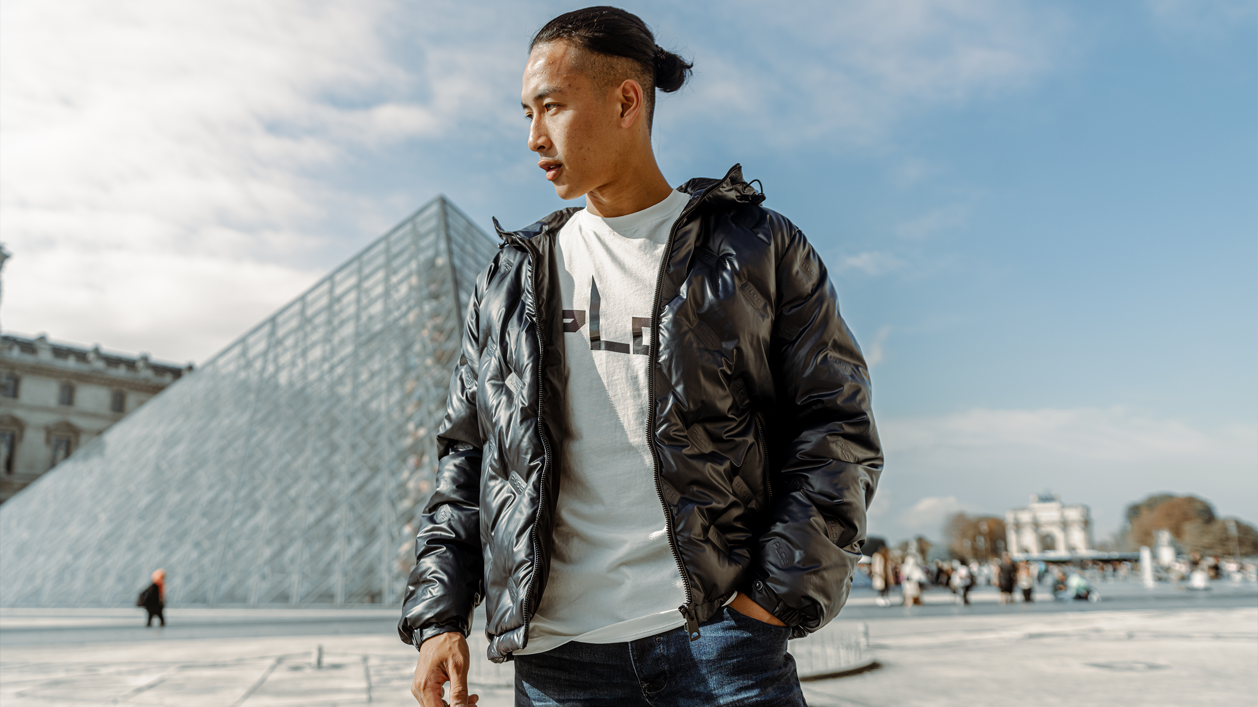 A young man with a fade haircut and long hair tied back, wearing a shiny black jacket and a white T-shirt, stands outdoors with a modern glass building and a cloudy sky in the background.