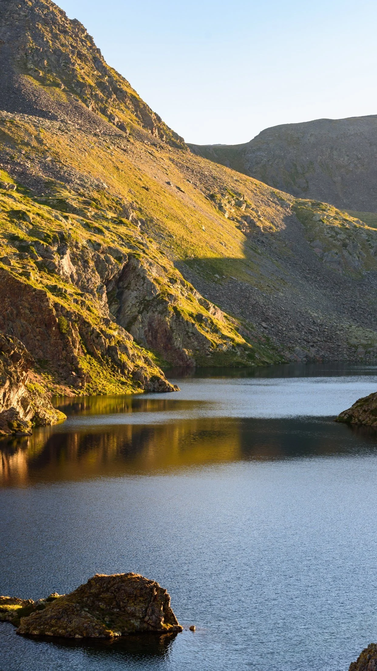 Scenic mountain landscape with steep rocky slopes covered in grass and a calm body of water reflecting the terrain.