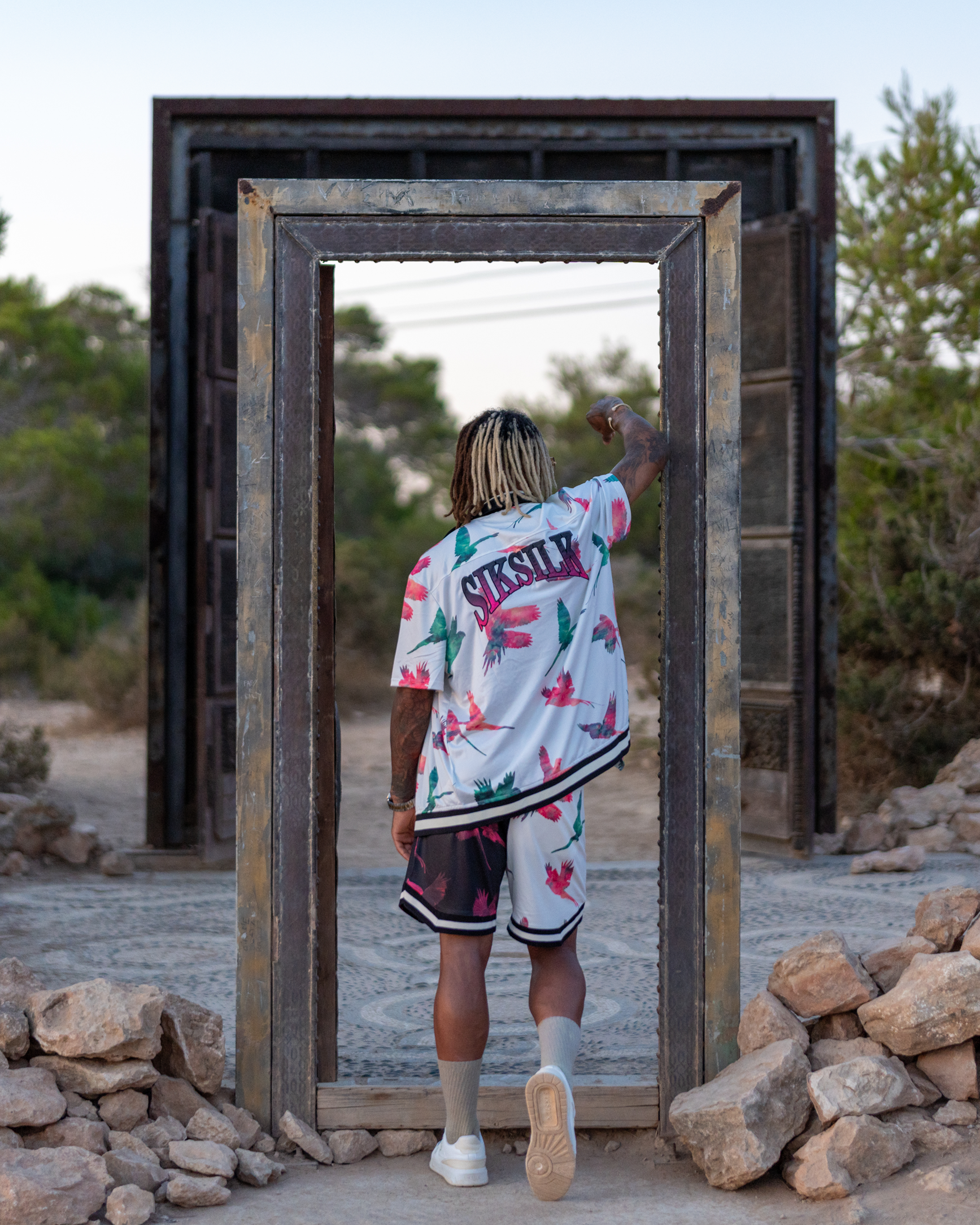 A person with dreadlocks wearing a colorful Hawaiian shirt and shorts, walking through a wooden frame in a rocky outdoor setting.