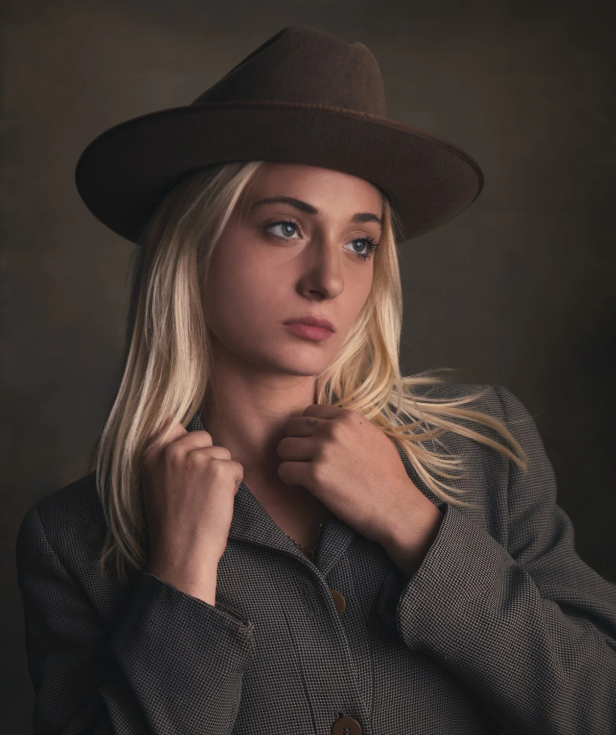 Editorial portrait of a blonde woman in a wide-brim felt hat and houndstooth coat against a mottled brown backdrop, Boston studio
