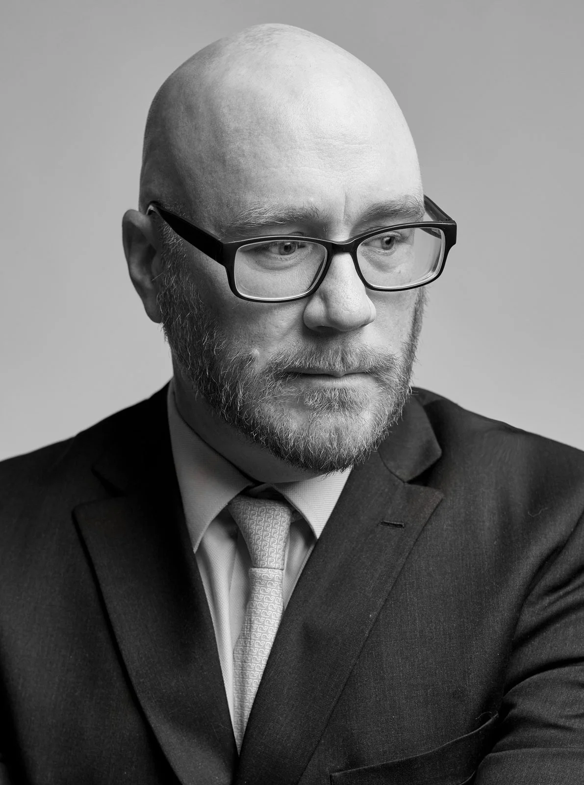 Black-and-white headshot of Kyle in dark-rimmed glasses, suit and tie, looking down against a gray background