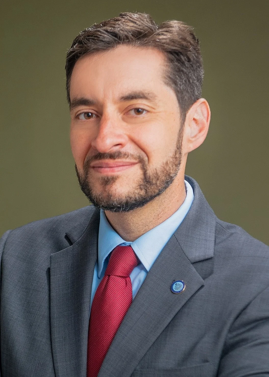 Headshot of a smiling bearded man in a gray suit, light blue shirt, red tie and lapel pin against an olive green backdrop