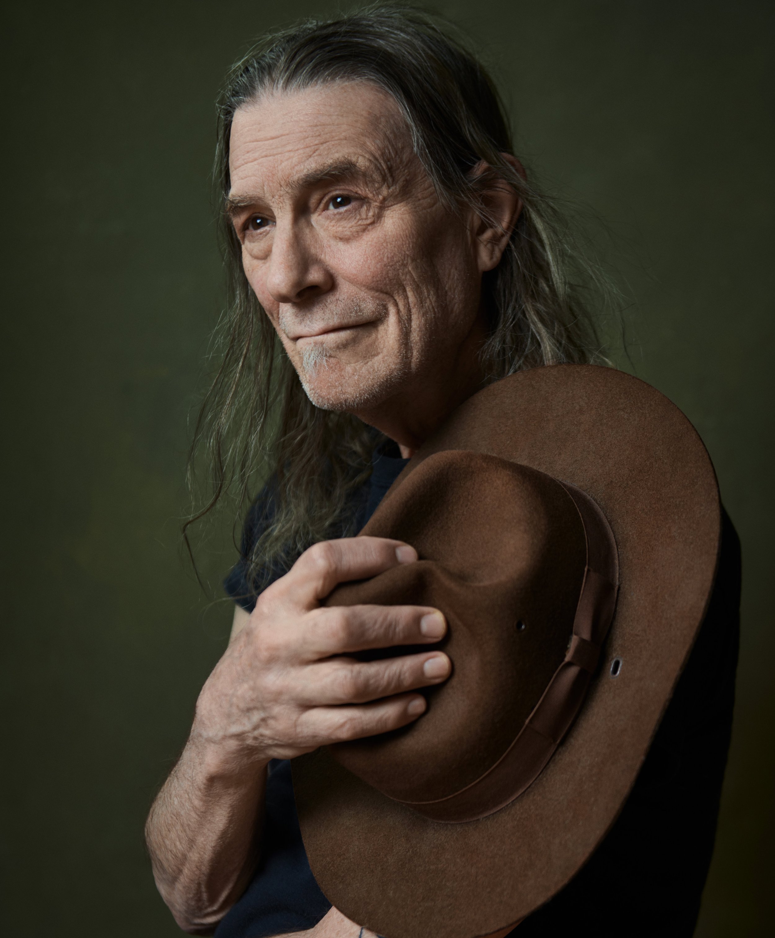 Boston musician Rick Berlin holding a felt hat, long grey hair, against a deep green painted backdrop