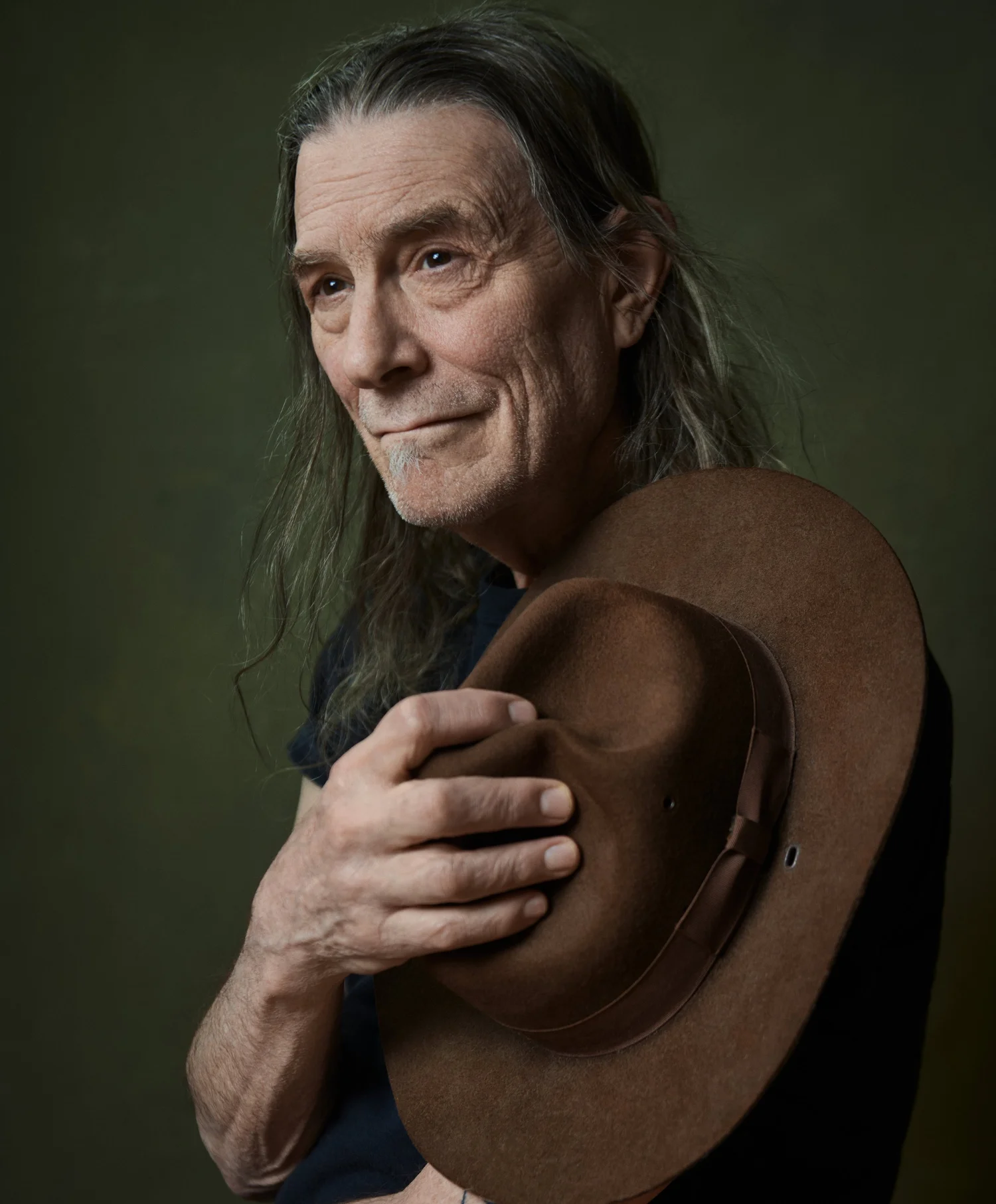 Color portrait of musician Rick Berlin, long gray hair, holding a brown felt hat to his chest against a dark green backdrop
