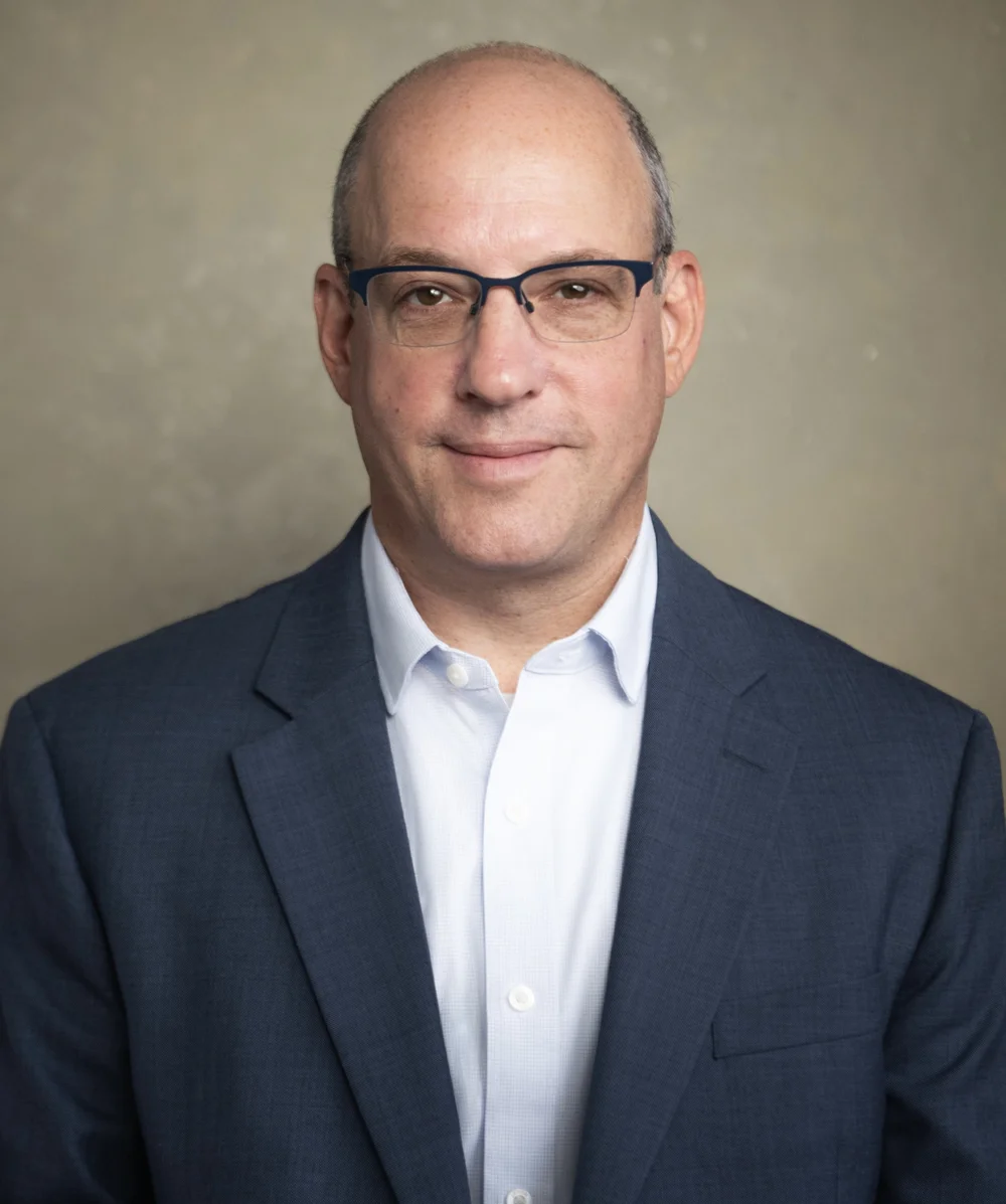 Headshot of a balding man with a slight smile in glasses, navy blazer and light blue shirt against a textured gray backdrop