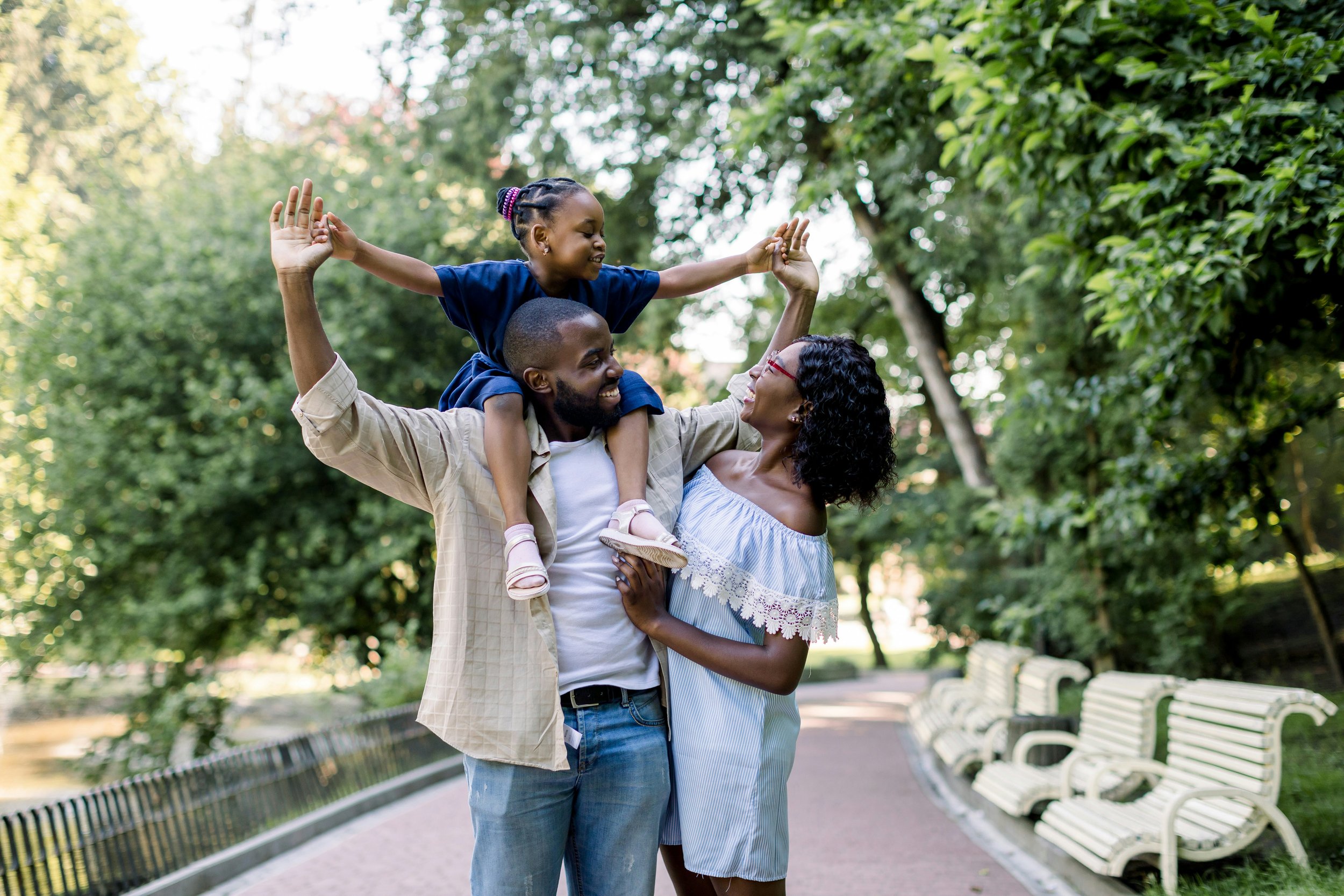 A family of three enjoying a moment outdoors in a park; father carries daughter on his shoulders, mother smiles at them, all are happy and smiling.