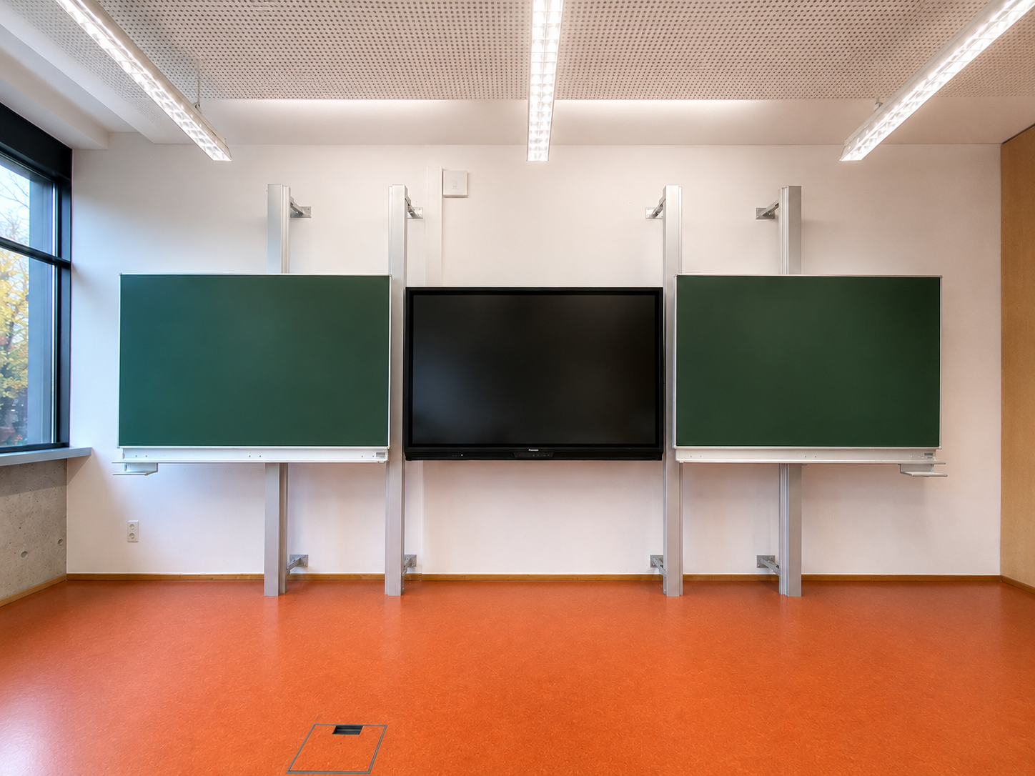 Leeres Klassenzimmer mit zwei grünen Tafeln, einem schwarzen Bildschirm in der Mitte, großen Fenstern auf der linken Seite und orangefarbener Bodenbelag.