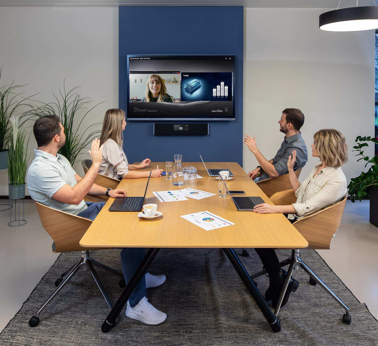 Fünf Personen sitzen in einem Konferenzraum um einen Holzkonferenztisch, während sie an einer Videokonferenz teilnehmen, mit Laptops, Papieren und Wassergläsern auf dem Tisch.