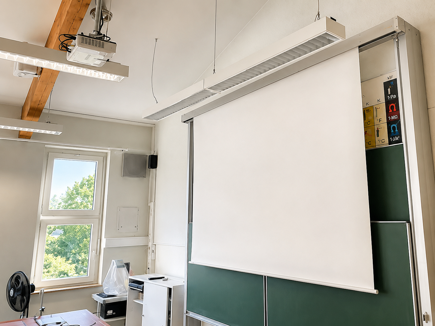 Leeres Klassenzimmer mit Tafel, Projektor, schwarzer Lampe, Fenster mit Blick nach draußen und verschiedenen technischen Geräten an der Wand.
