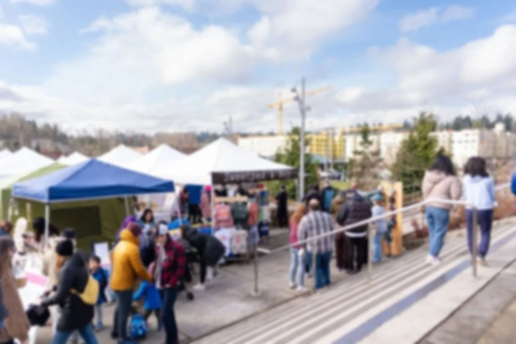 Blurred outdoor scene of people in line at a market or outdoor event with tents, a crane in the background, and cloudy sky.