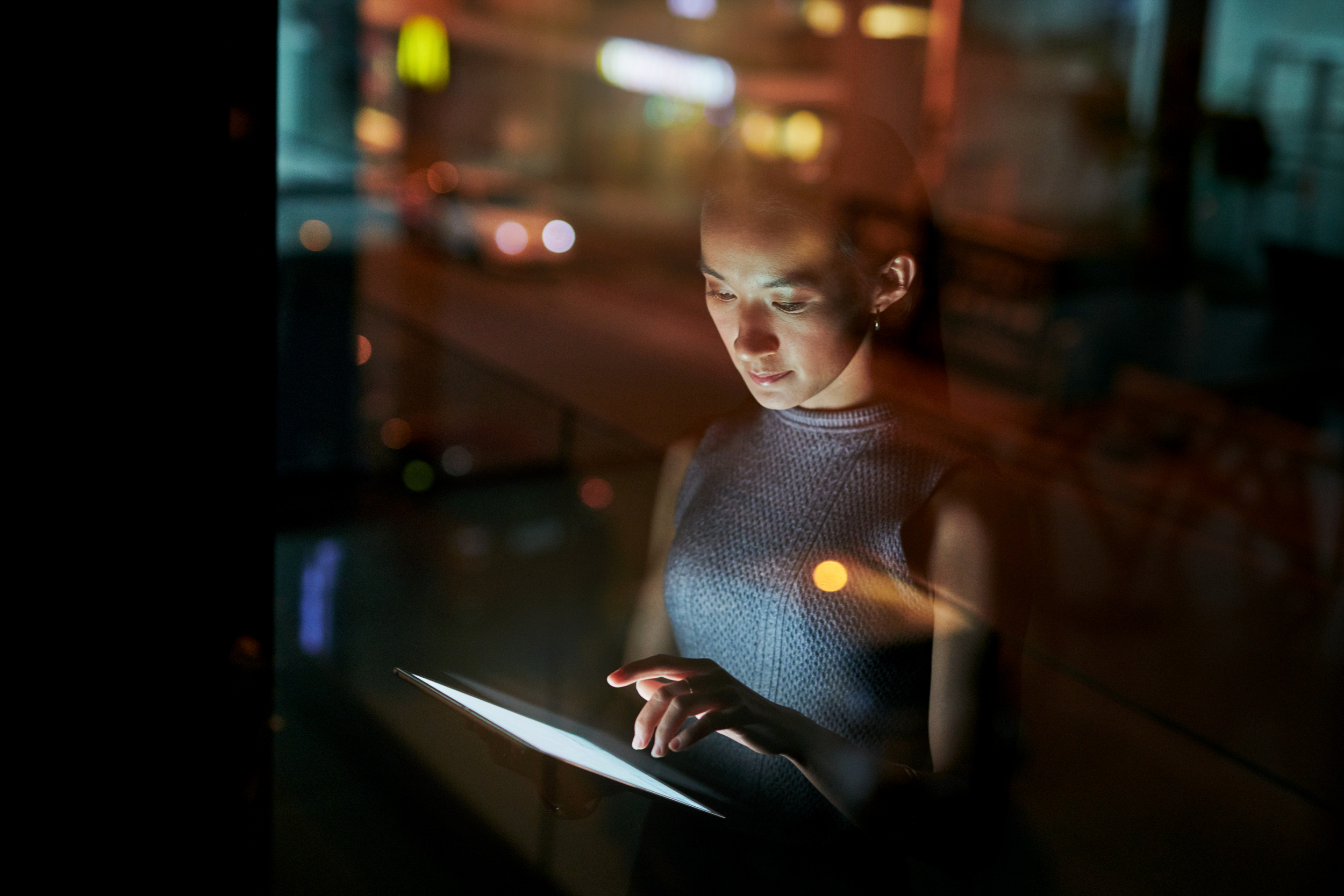 A woman looking at a smartphone screen at night, reflected on a glass surface with city lights blurred in the background.