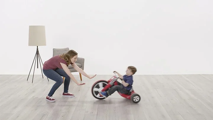 Woman playing with young boy on a small red ride-on toy in a minimalistic room with light-colored wooden floor, a gray chair, and a tall floor lamp.
