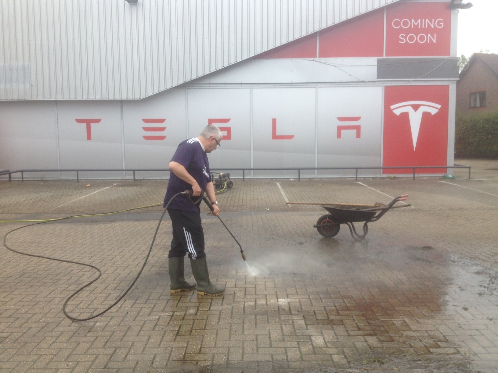 A person is power washing a paved area in front of a building with a Tesla logo and 'Coming Soon' sign. A wheelbarrow is visible nearby.