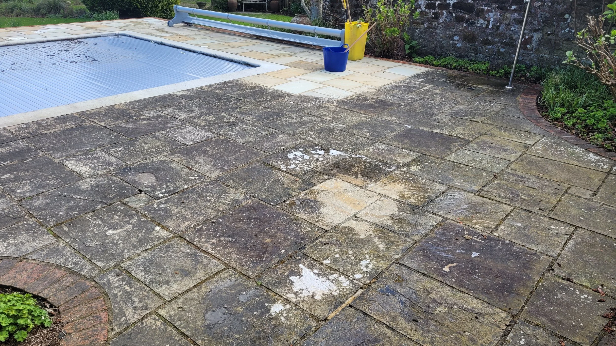 Stone patio with wet patches near a covered swimming pool, cleaning supplies including a mop and bucket, and garden area.