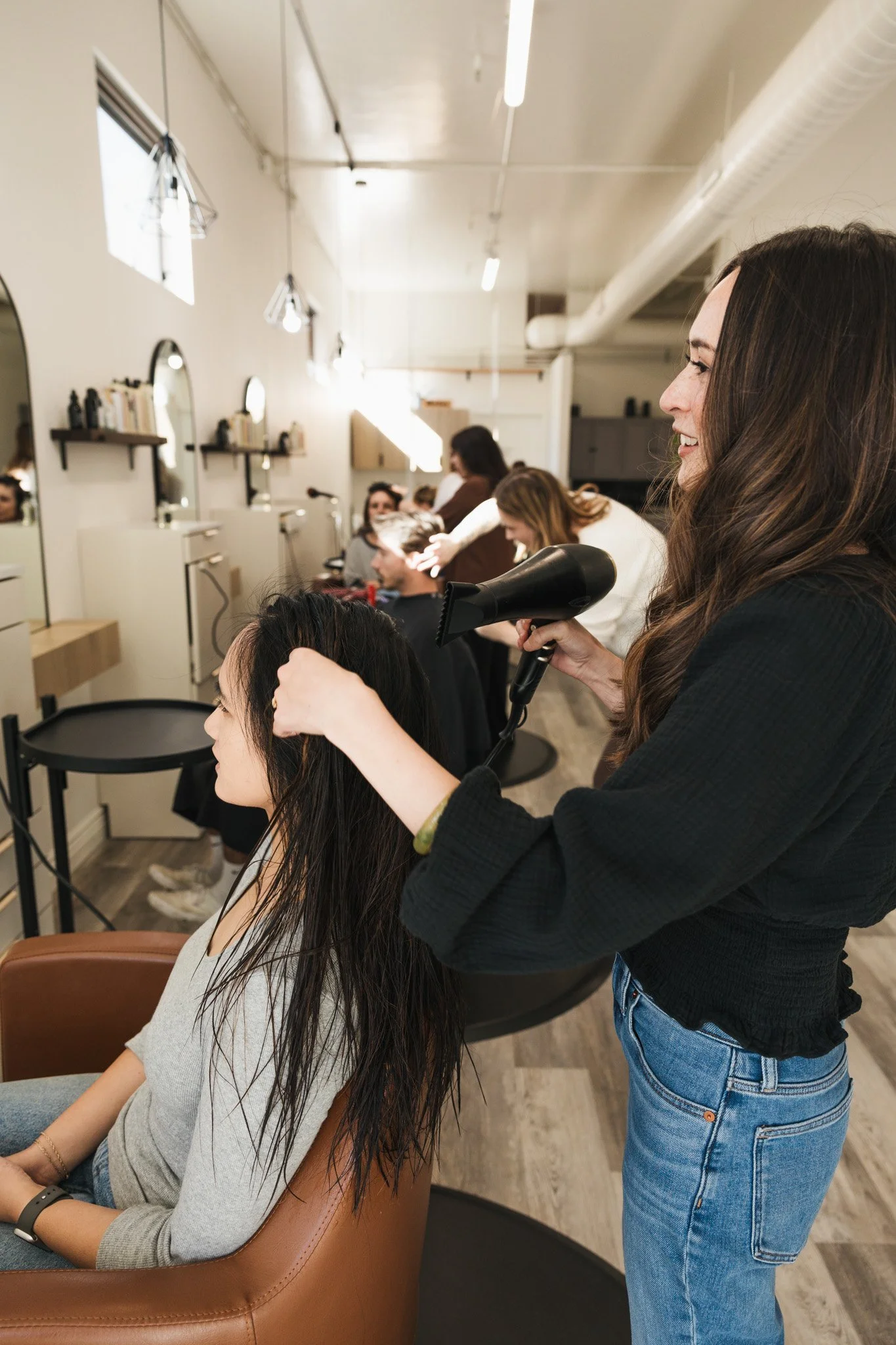 A hairstylist drying a woman's hair in a salon with other customers getting hair treatments. The salon has a modern decor with mirrors, shelves, and several overhead lights.