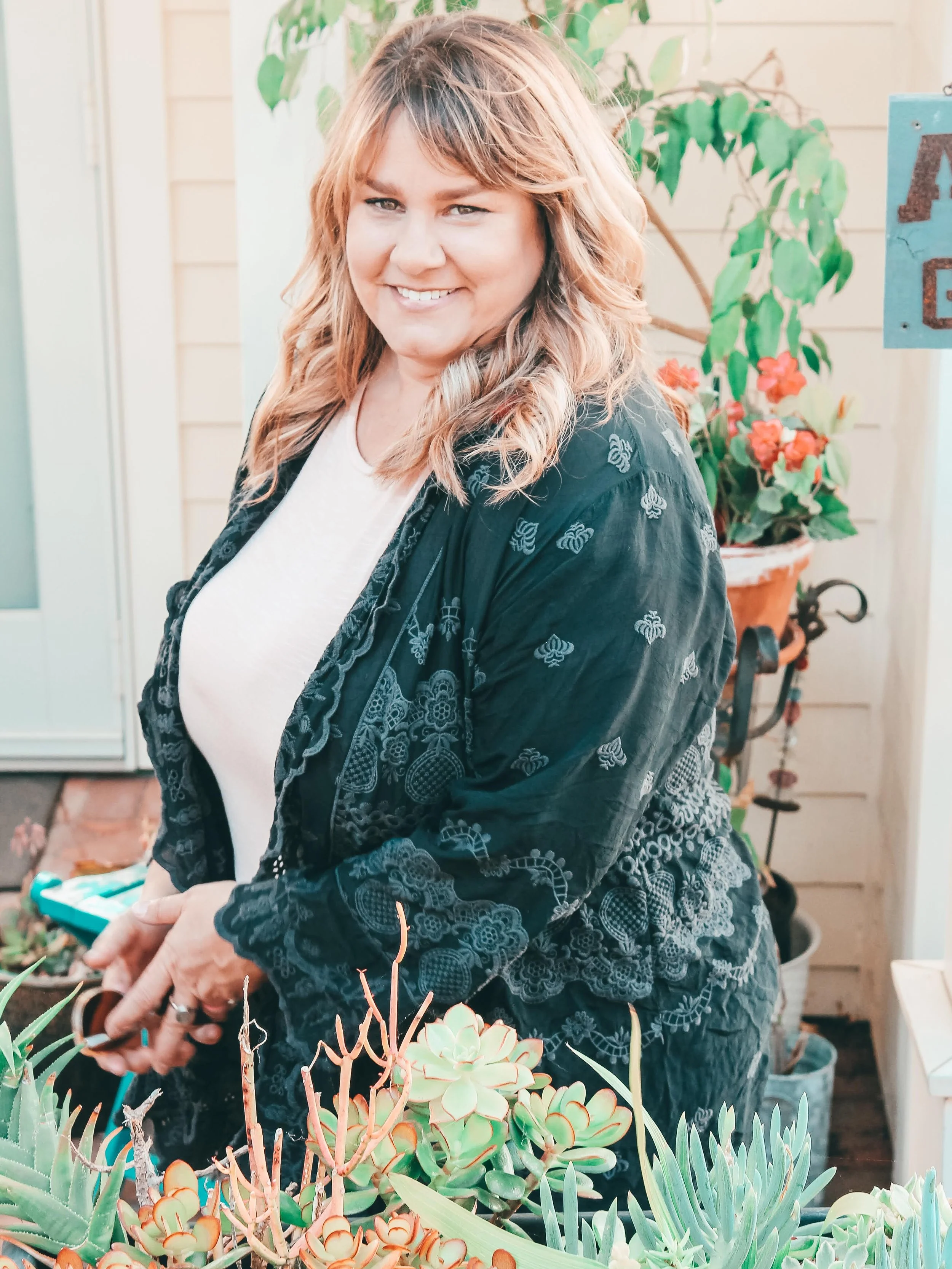 A woman with light brown, wavy hair smiling outdoors, surrounded by various potted succulent plants, with a beige wooden wall and green plant in the background.