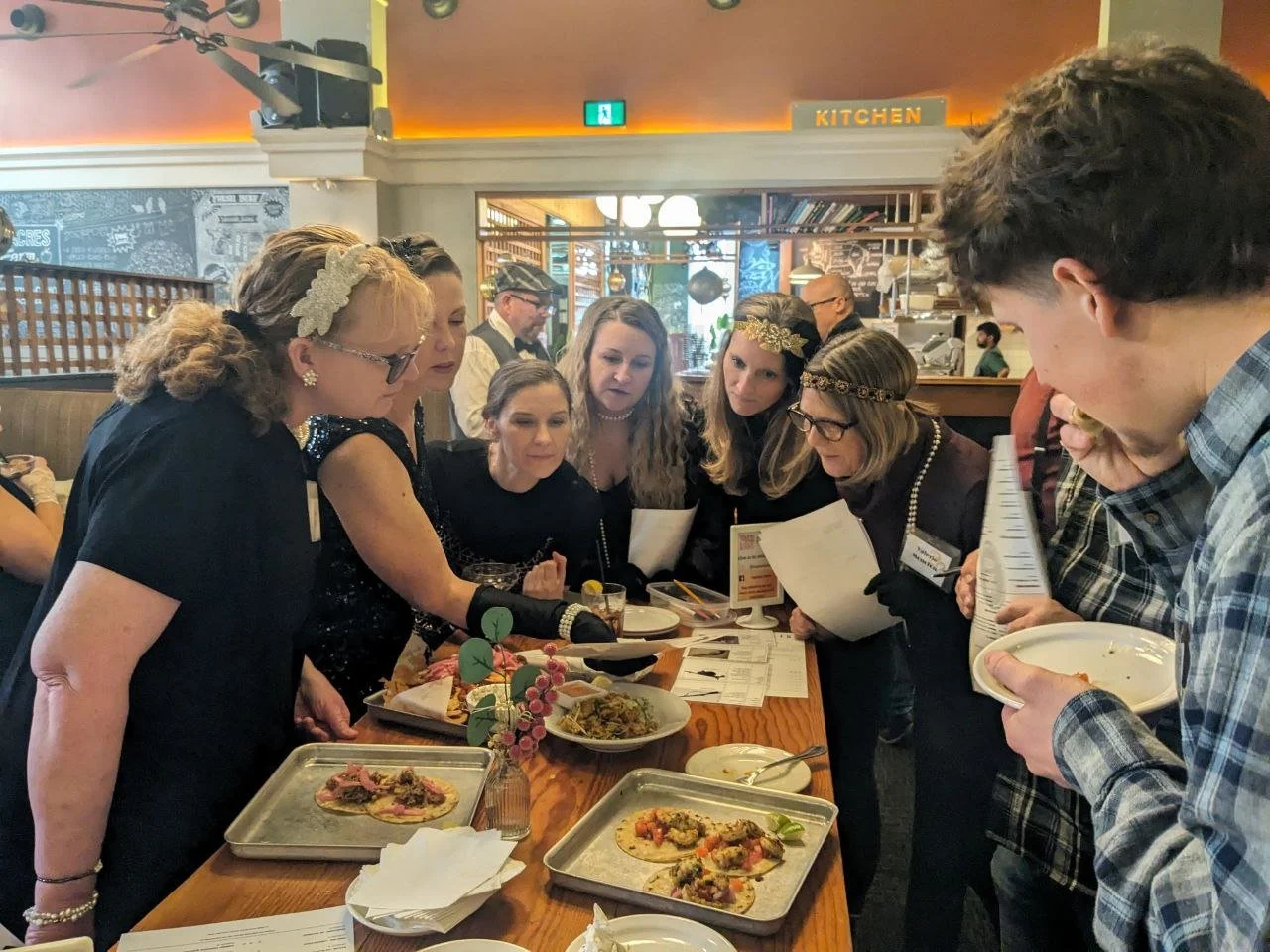 A group of 7 people are around table which has tacos and printed material on. the group are all looking at one piece of paper intently.  Six of the people in the group are dressed up in 1920's style costumes.