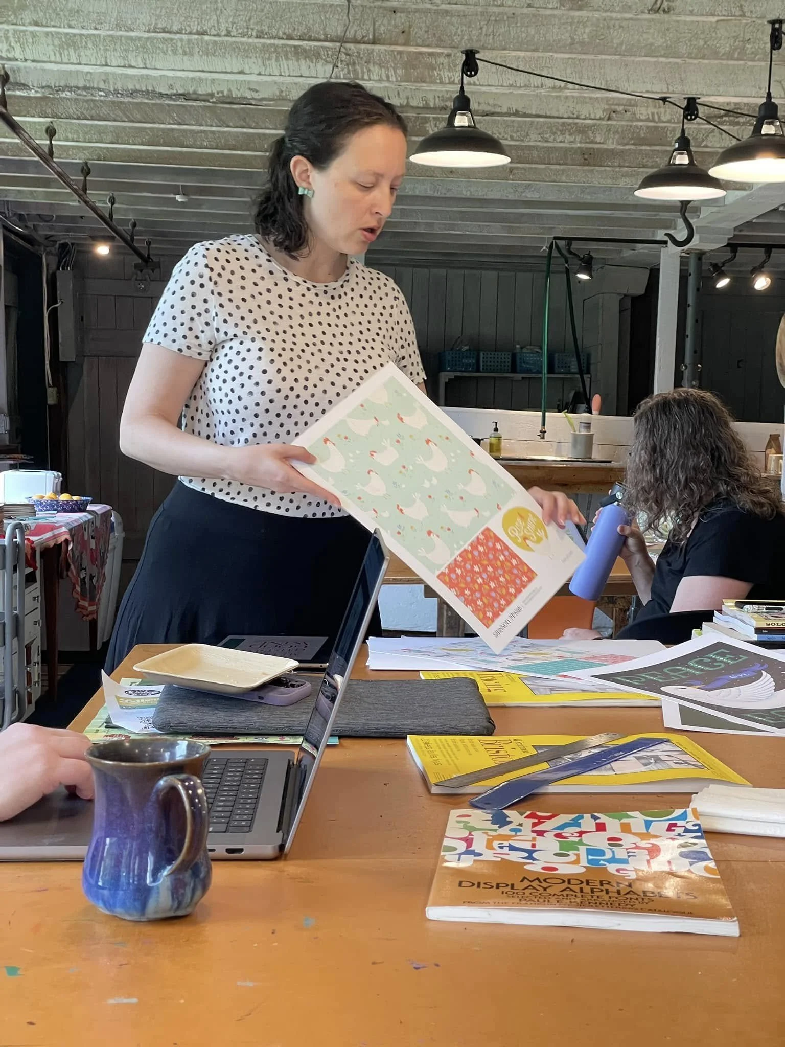 Instructor holding up a printed pattern design sample while presenting at the long studio table in the barn