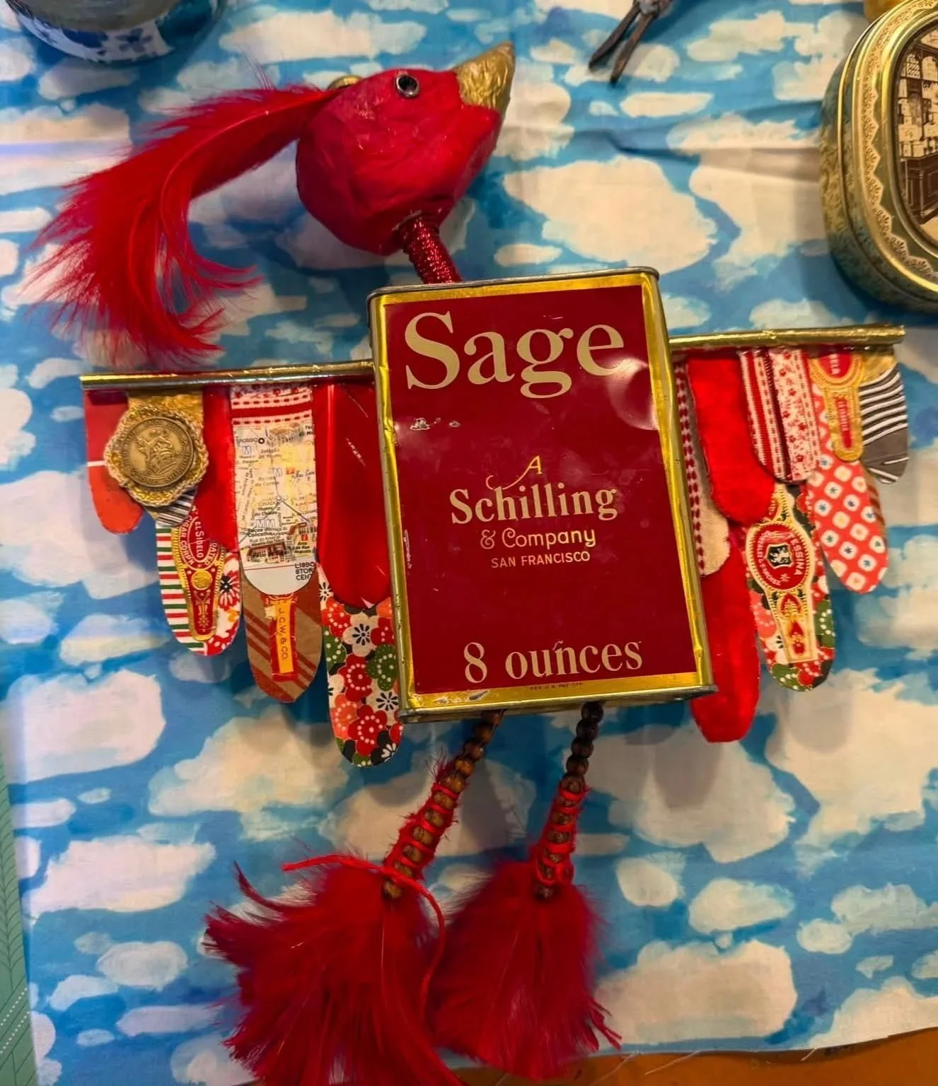 Mixed media assemblage with a red feathered bird, vintage Sage box and tassels on cloud-patterned fabric