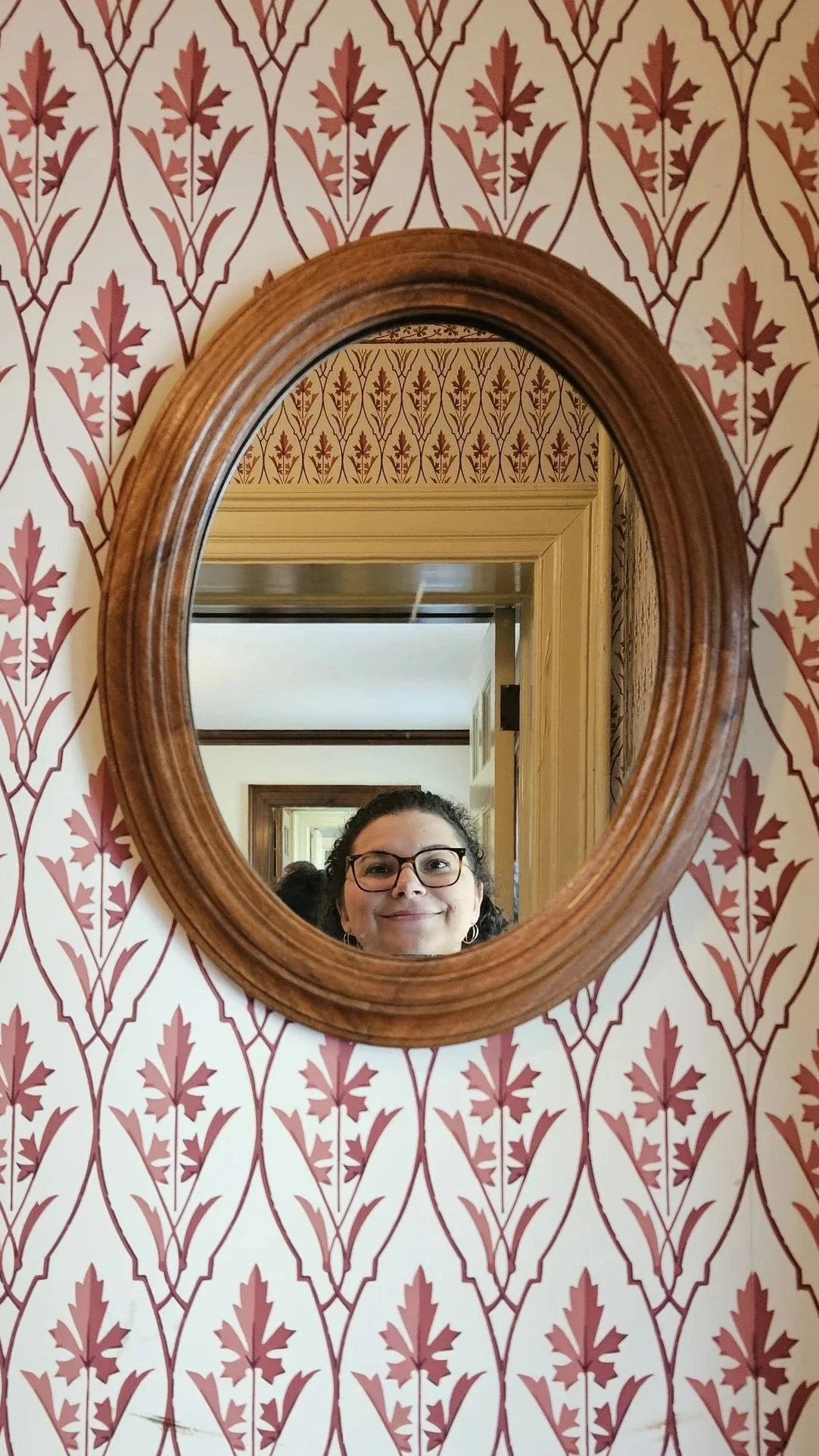Oval wooden mirror on red leaf patterned wallpaper reflecting a smiling artist