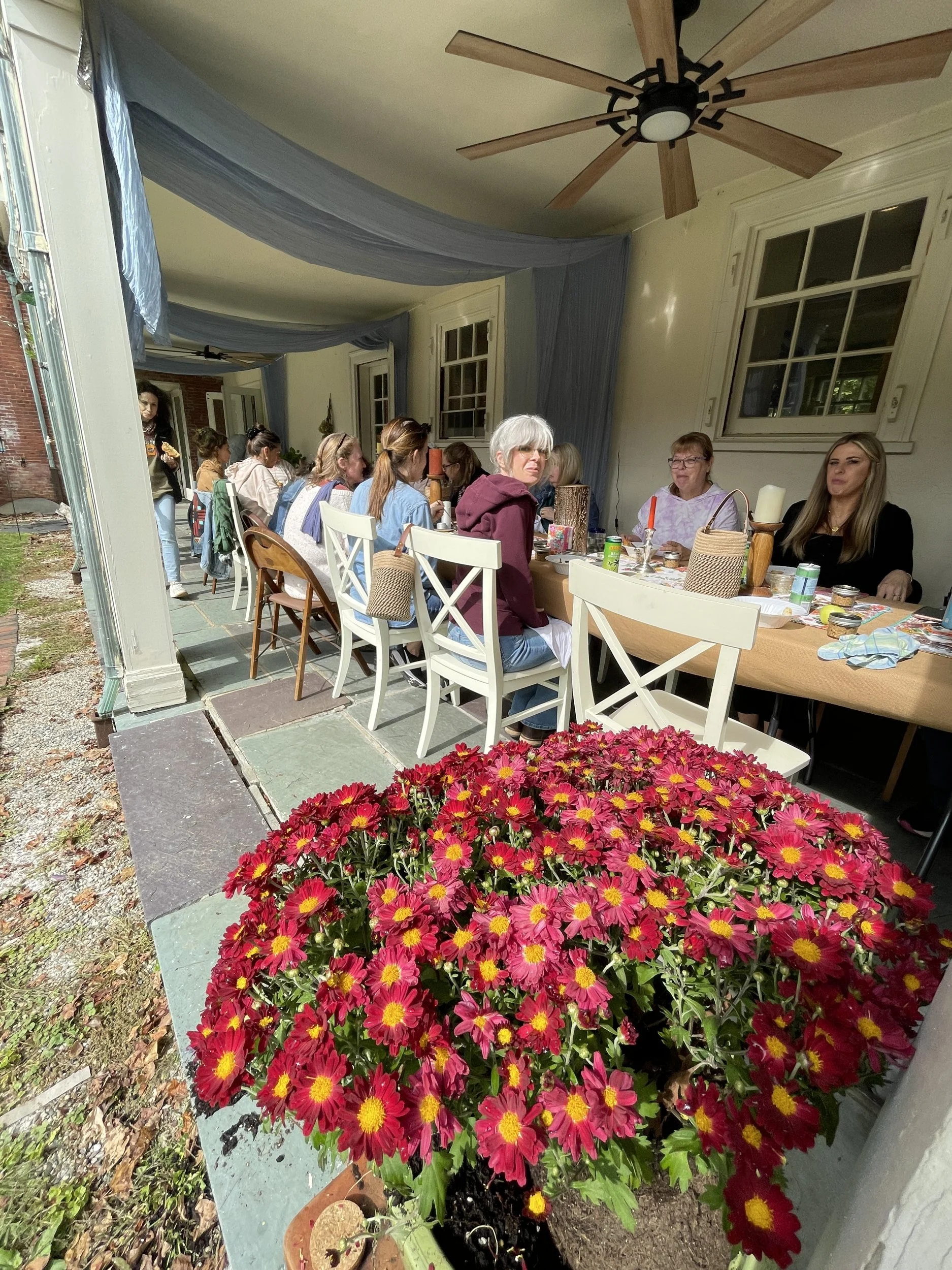 Artists seated at a long table on the covered porch behind a large pot of red chrysanthemums