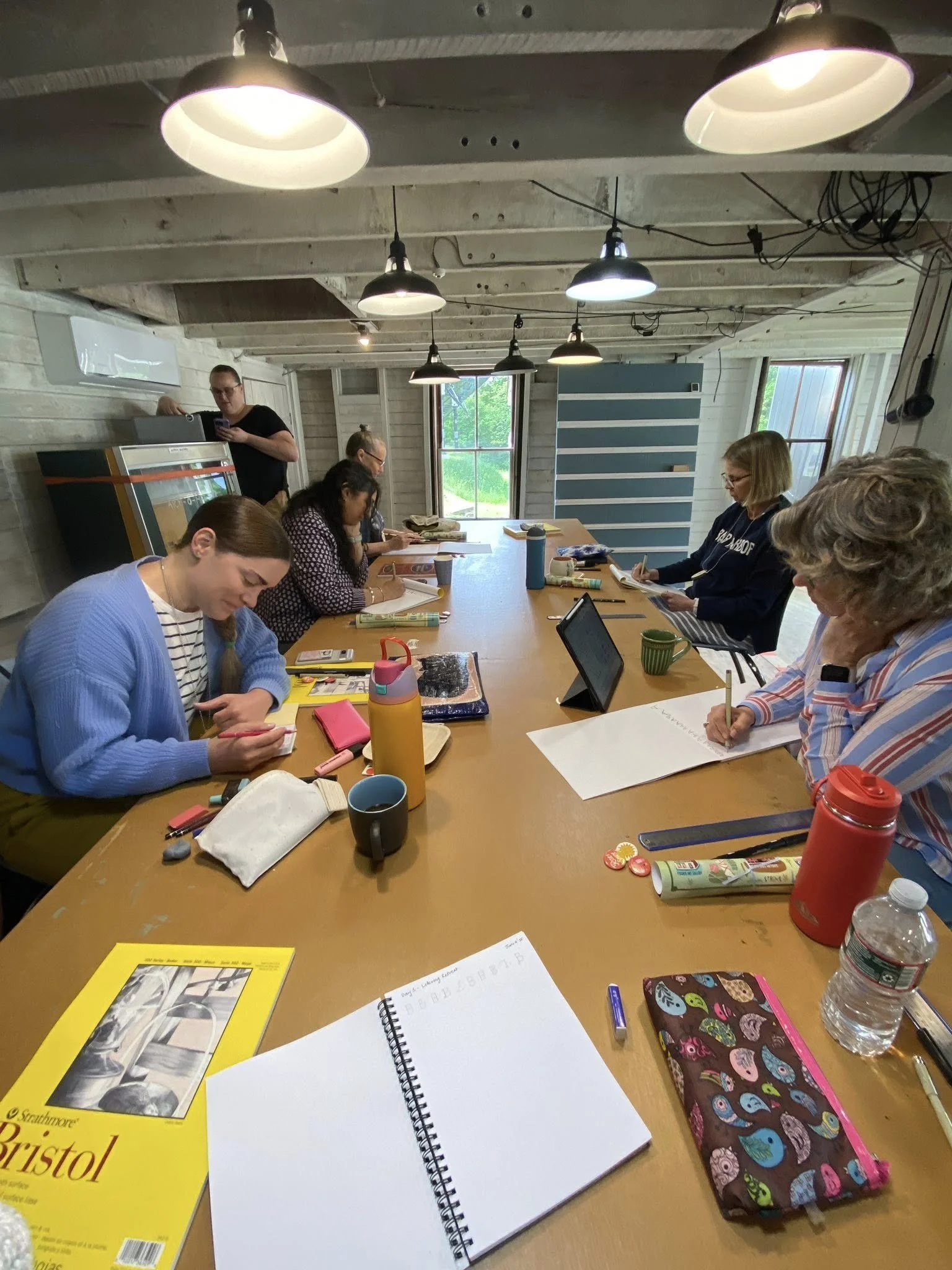 Artists drawing and lettering at the long studio table in the barn with Bristol pads, sketchbooks and colorful pencil pouches