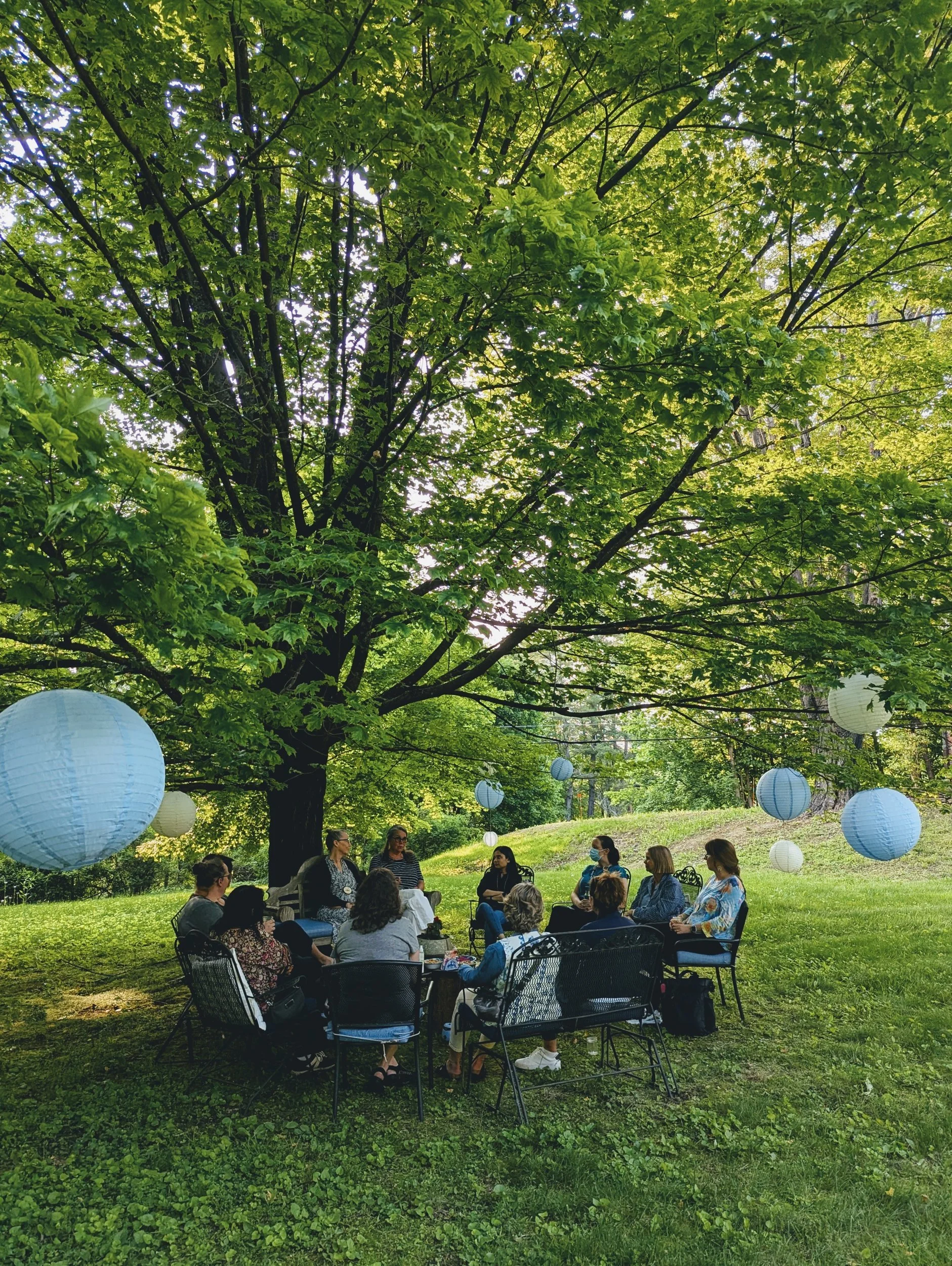 Retreat artists gathered in a circle of chairs on the lawn under a large maple tree hung with blue paper lanterns