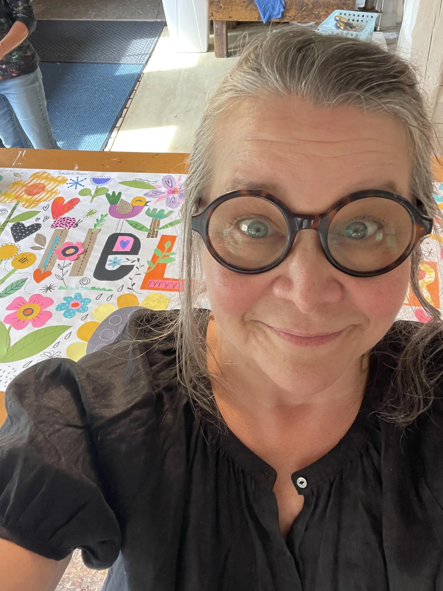 Jennifer Nelson in round glasses taking a close selfie over the Hello Spring collage laid on the table.