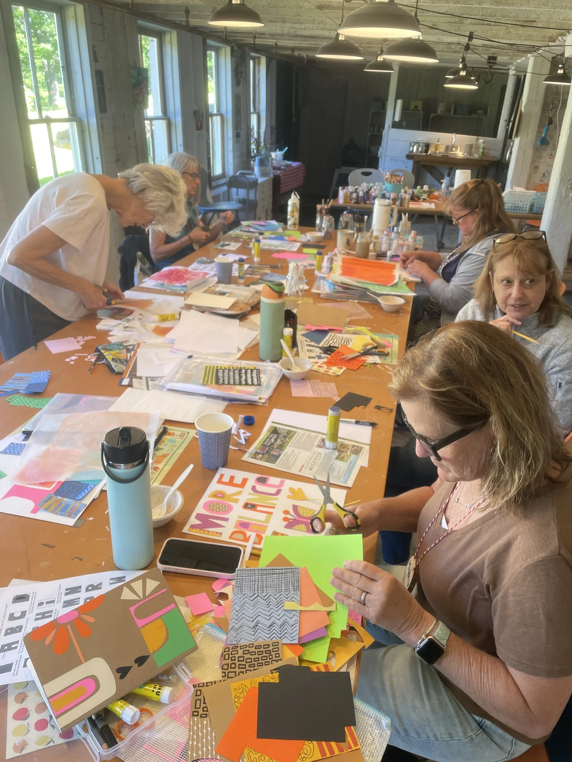 Artists cutting colorful papers and assembling collages at a long worktable in the barn studio.