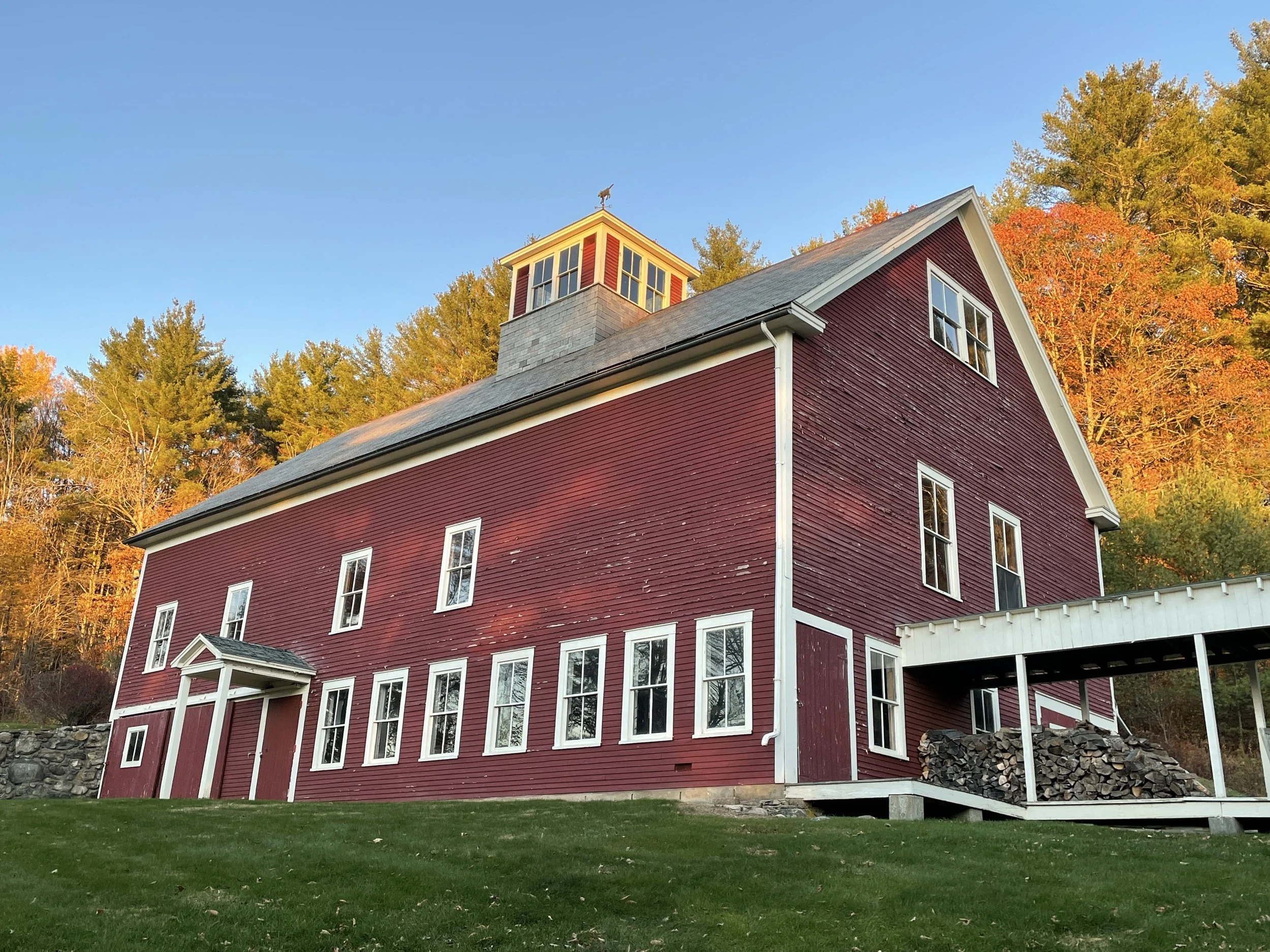 Red barn art studio with a cupola and white porch in golden light with fall foliage behind it