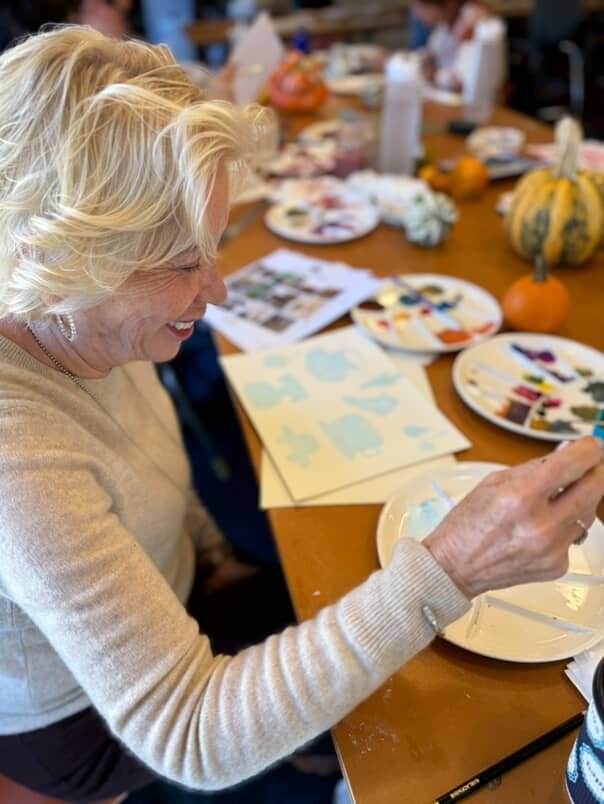 Smiling artist painting with watercolors at a long table decorated with small pumpkins and gourds