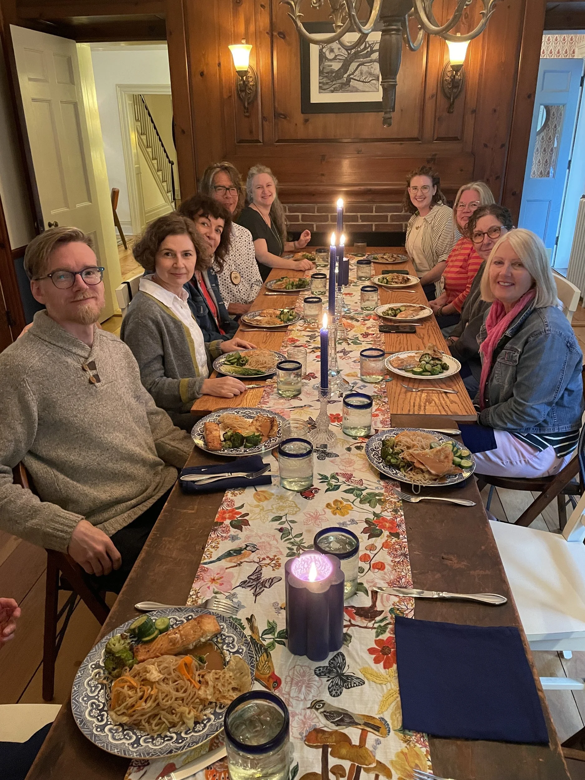 Retreat artists smiling around a long wooden dining table set with plates of salmon and noodles, blue candles and a bird and butterfly table runner