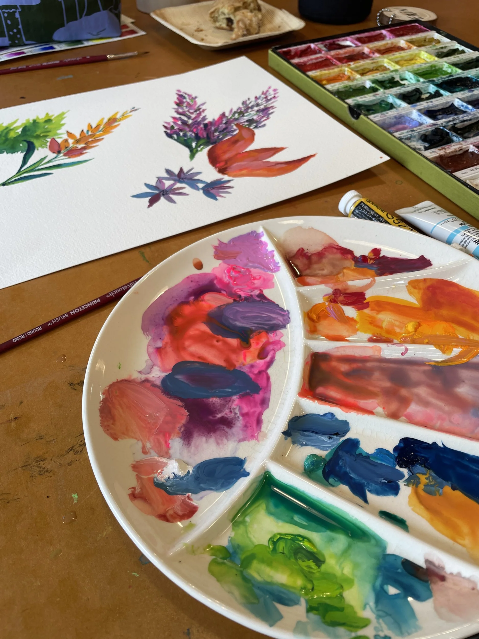 Watercolor lupine studies on paper beside a white palette mixed with bright pinks, blues and greens
