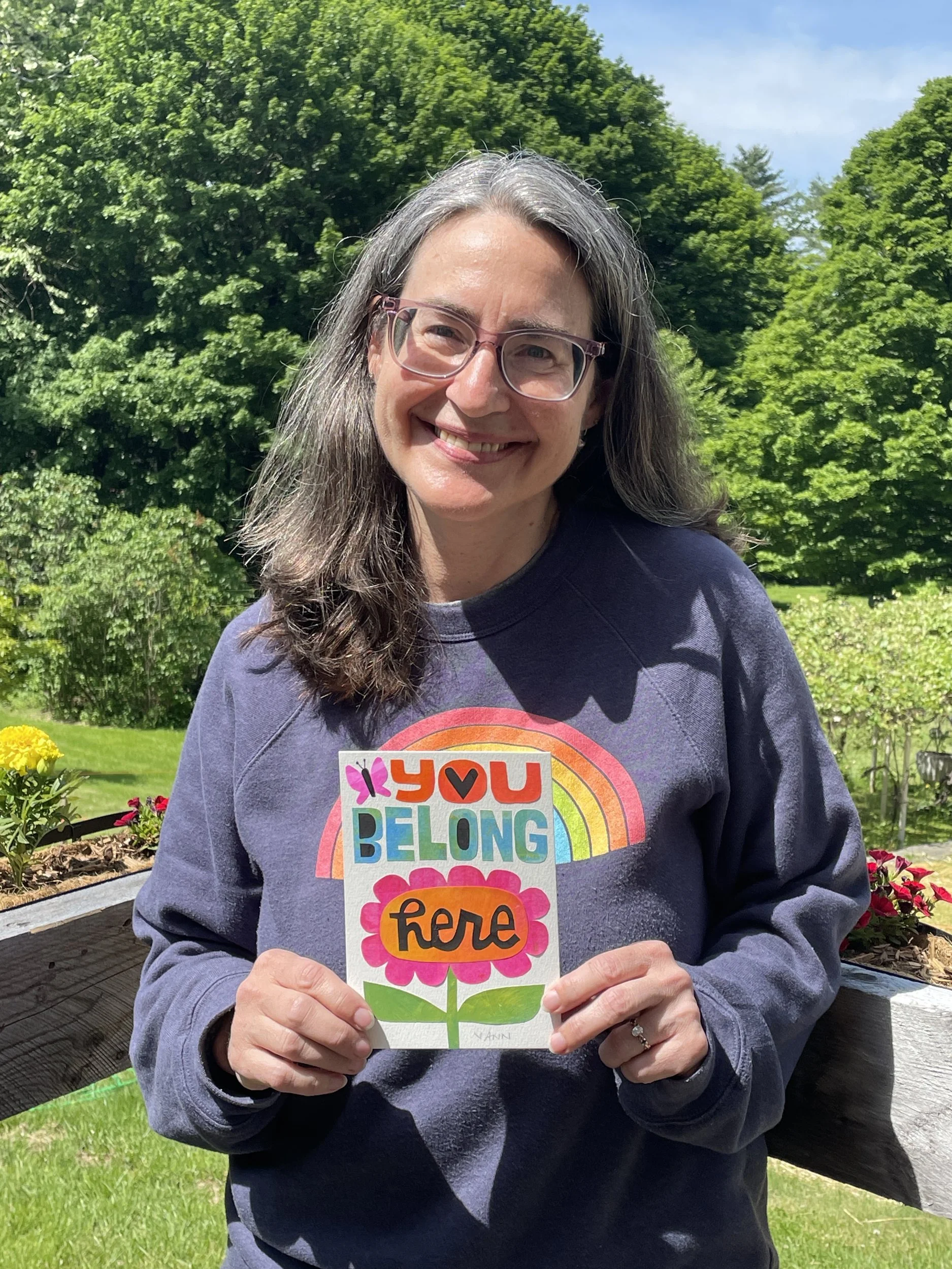 Smiling artist outdoors holding a lettered card that reads You Belong Here with a rainbow and pink flower.