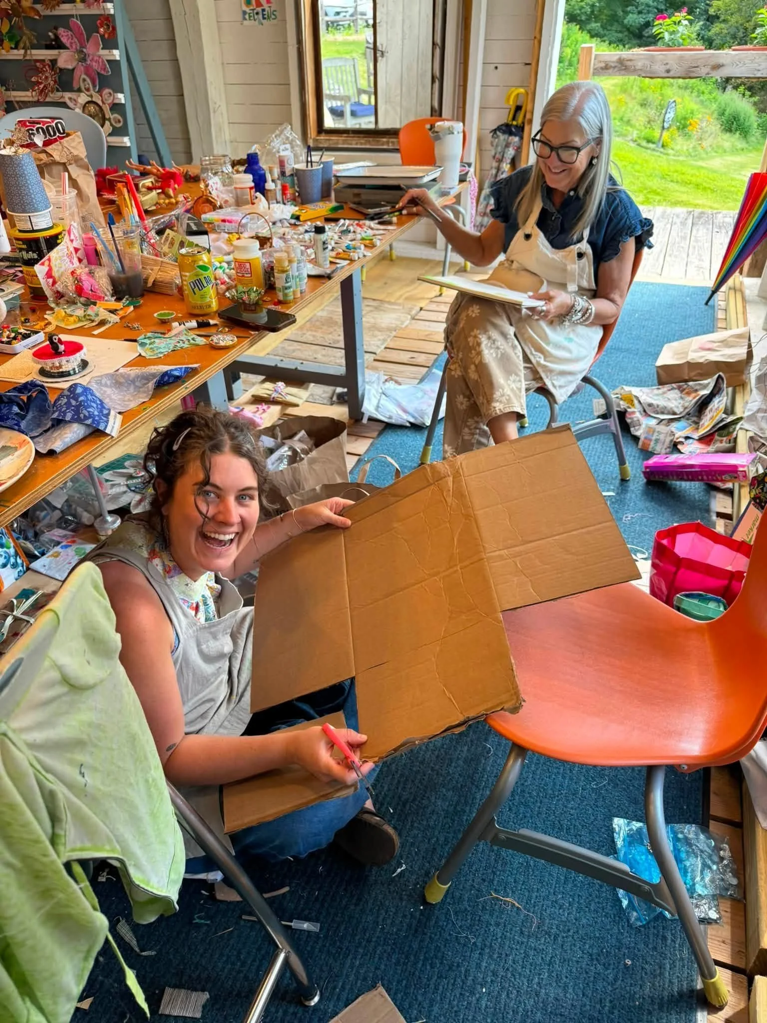 Busy barn studio filled with art supplies as an artist opens a cardboard box while another works at a table