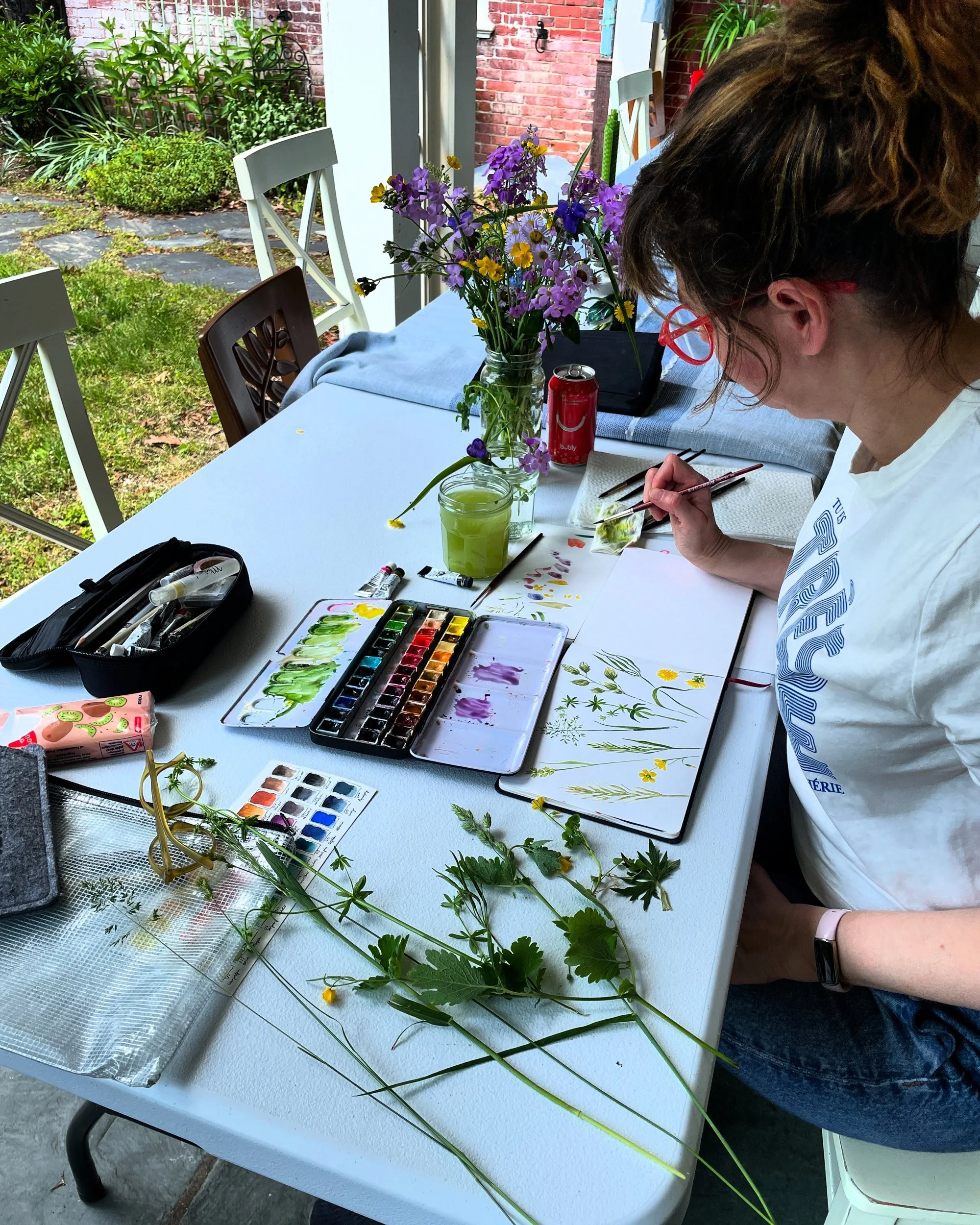 Artist painting a watercolor of wildflowers at a porch table with fresh-picked flowers, a paint palette and a water jar