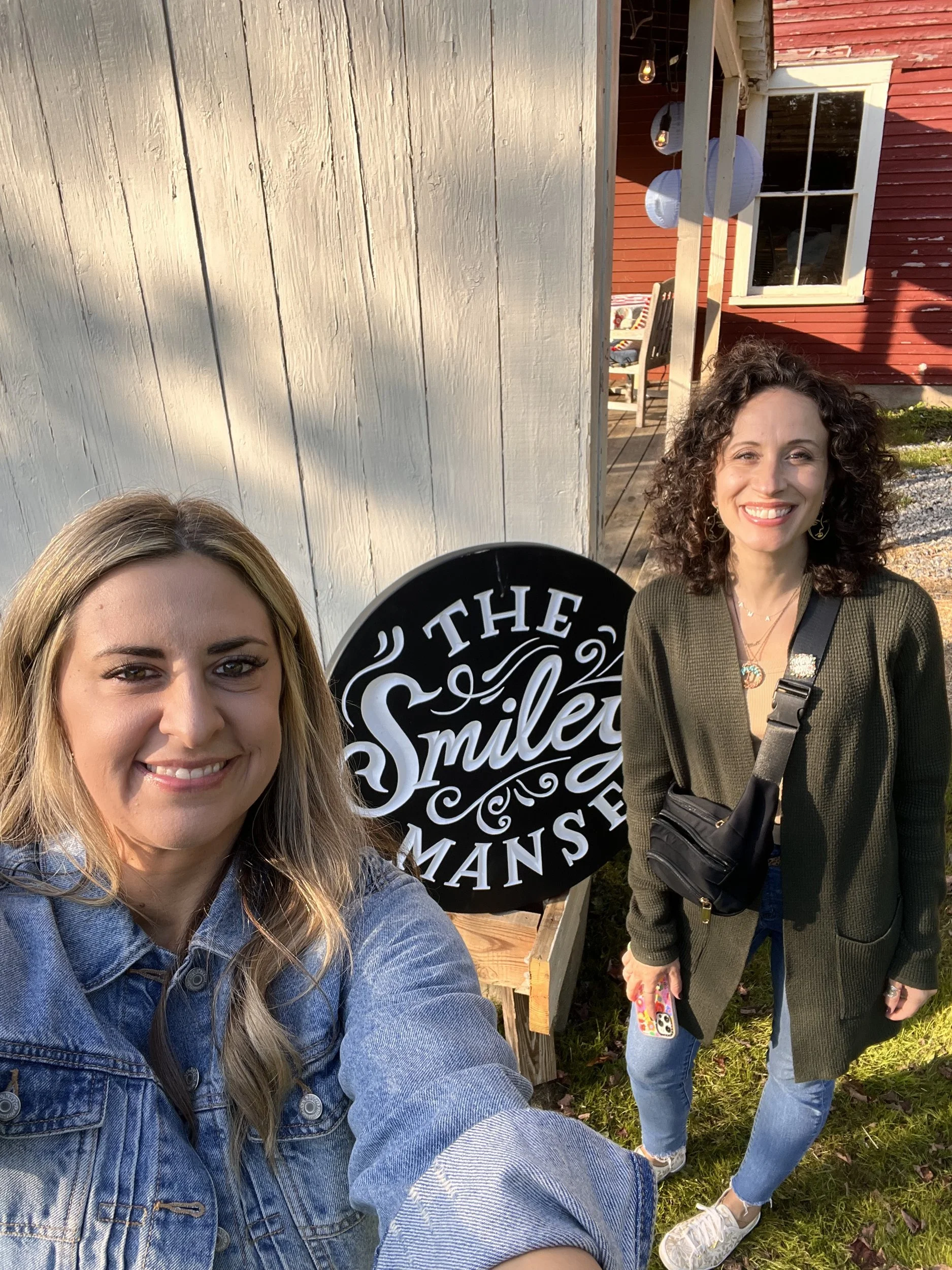 Two smiling artists posing beside the round Smiley Manse sign near the barn